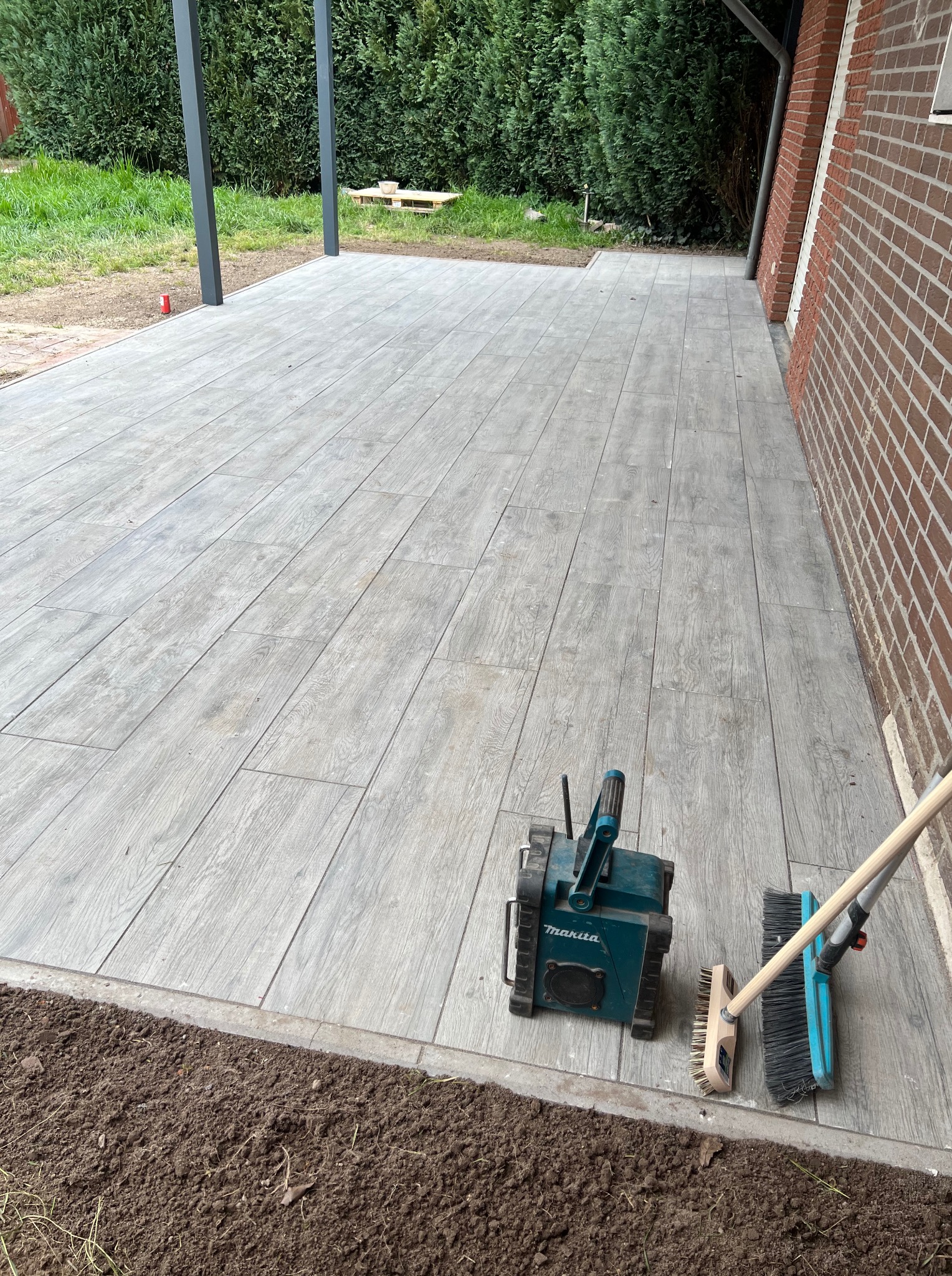 Nowo ułożona, drewnopodobna, szara kostka brukowa na tarasie, z widocznymi narzędziami pracy (szczotki, radio) i świeżo przygotowanym obrzeżem pod nasadzenia.