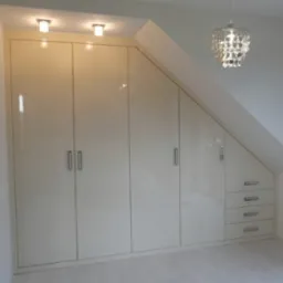 Biała szafa wnękowa na poddaszu ze srebrnymi uchwytami i czterema szufladami, oświetlona lampami sufitowymi i wiszącym, dekoracyjnym żyrandolem.