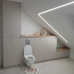 Beżowa zabudowa meblowa w łazience na poddaszu ze skośnym sufitem i oświetleniem LED. Wisząca toaleta z dywanikiem. Minimalistyczny styl.