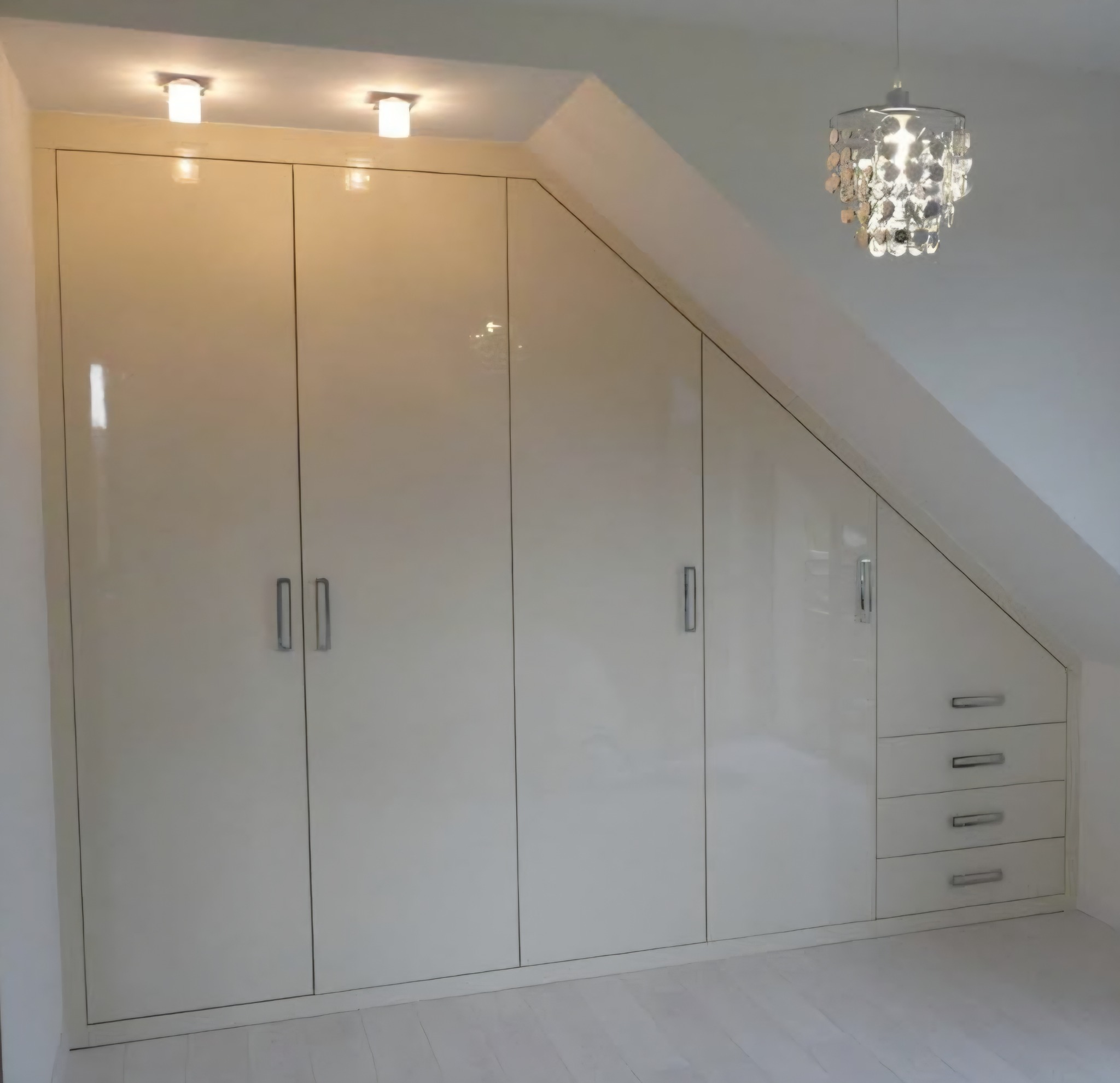 Biała szafa wnękowa na poddaszu ze srebrnymi uchwytami i czterema szufladami, oświetlona lampami sufitowymi i wiszącym, dekoracyjnym żyrandolem.