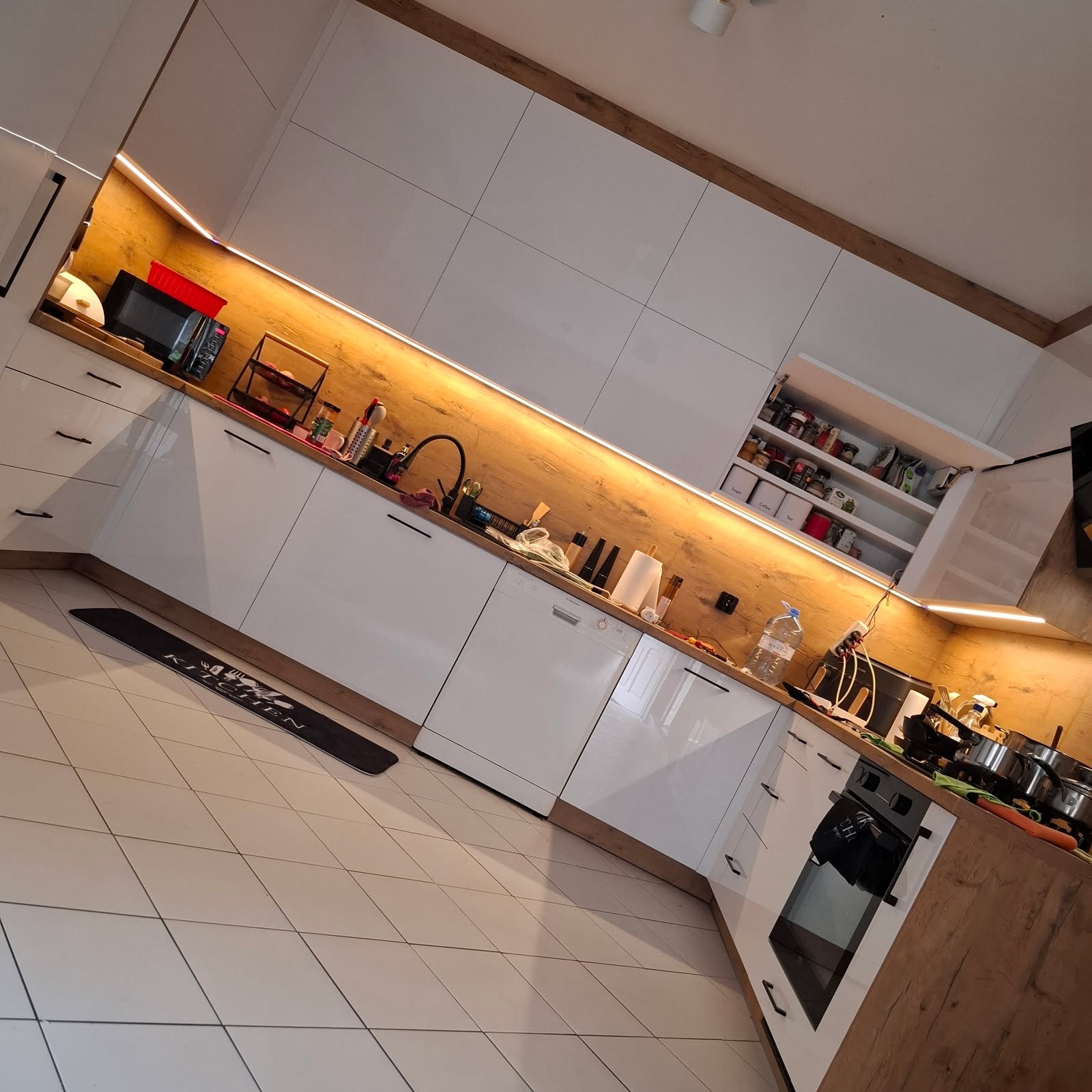 Nowoczesna kuchnia z białymi, błyszczącymi frontami i drewnopodobnym blatem, oświetlona listwami LED. Na blacie akcesoria kuchenne, na podłodze dywanik.