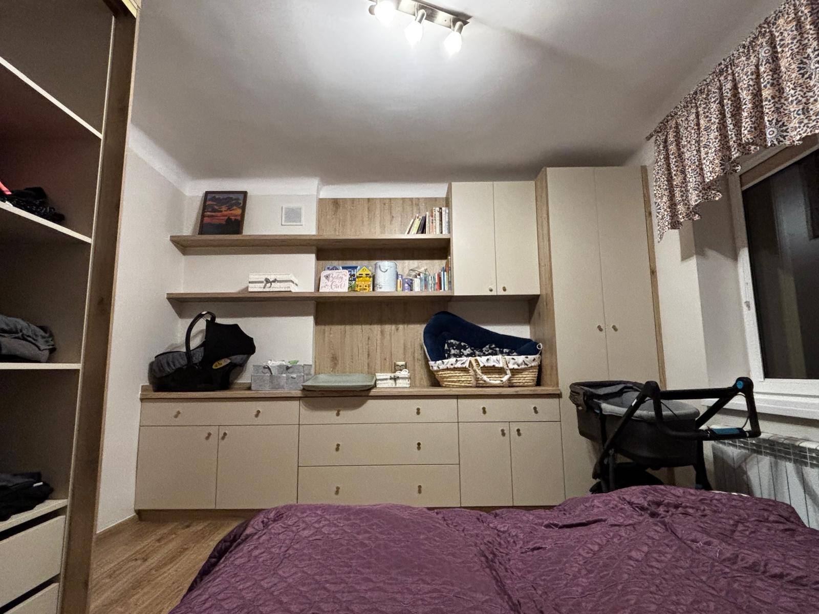 Beżowa szafa wnękowa z półkami w pokoju dziecięcym. Na blacie szafy akcesoria dla niemowląt. Widok z perspektywy łóżka z fioletową narzutą.