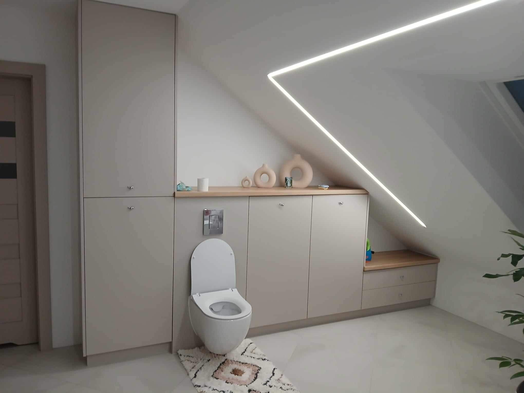 Beżowa zabudowa meblowa w łazience na poddaszu ze skośnym sufitem i oświetleniem LED. Wisząca toaleta z dywanikiem. Minimalistyczny styl.