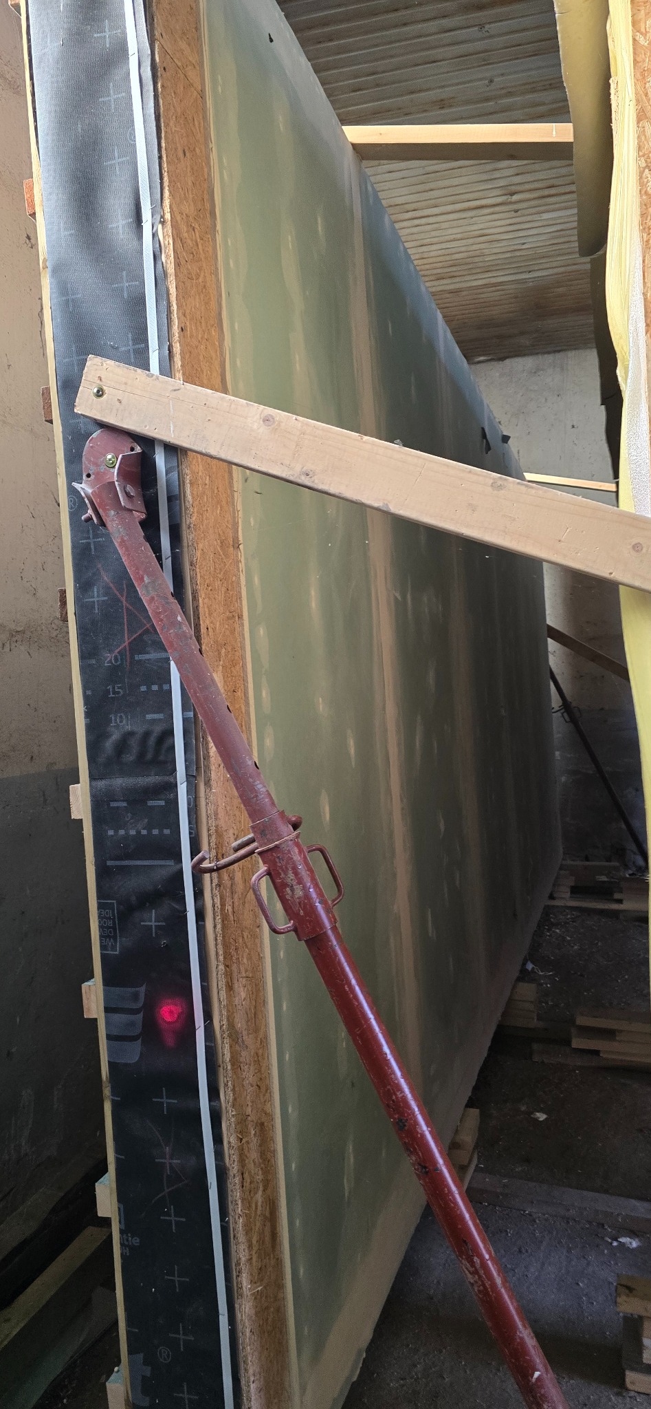 Wznoszenie ściany z płyty warstwowej, podpartej stemplem budowlanym. Widoczne warstwy izolacji i konstrukcji. W tle surowy strop budynku w trakcie budowy.