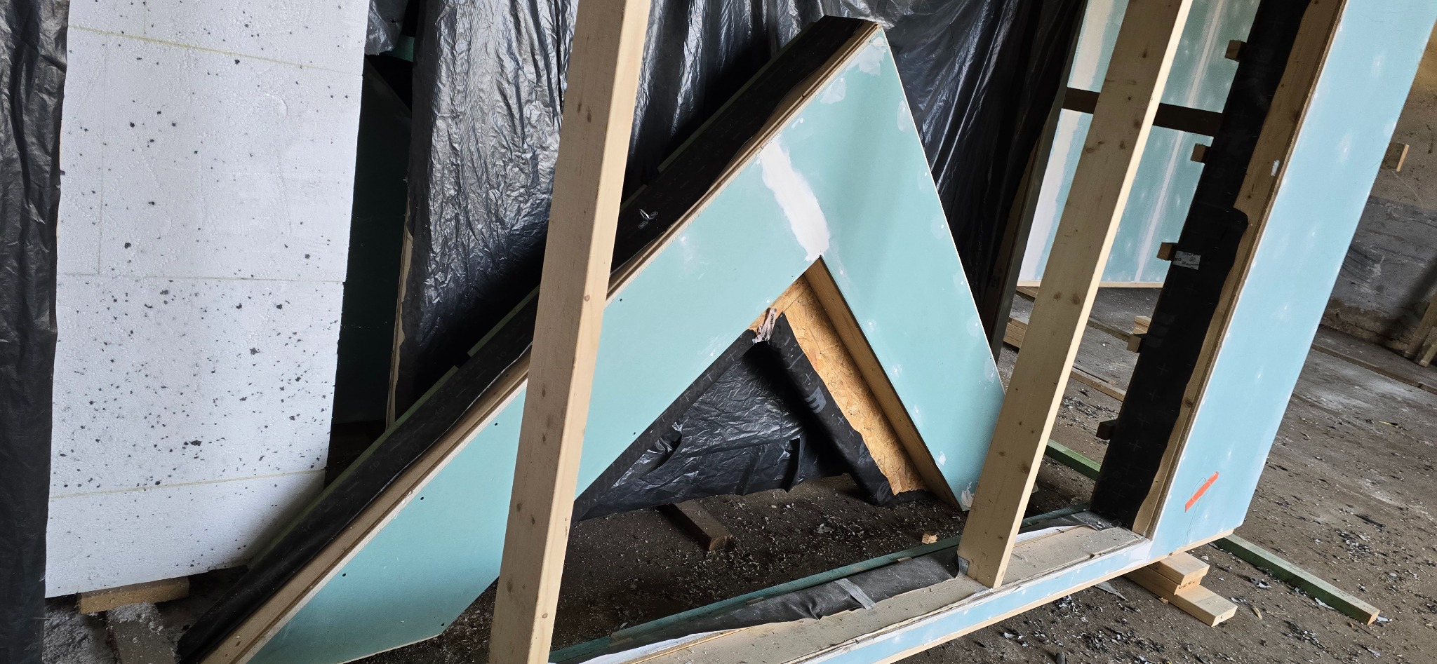 Konstrukcja ściany szkieletowej z płyt gipsowo-kartonowych i izolacją, widoczne drewniane elementy ramy i czarna folia izolacyjna. Etap budowy domu szkieletowego.