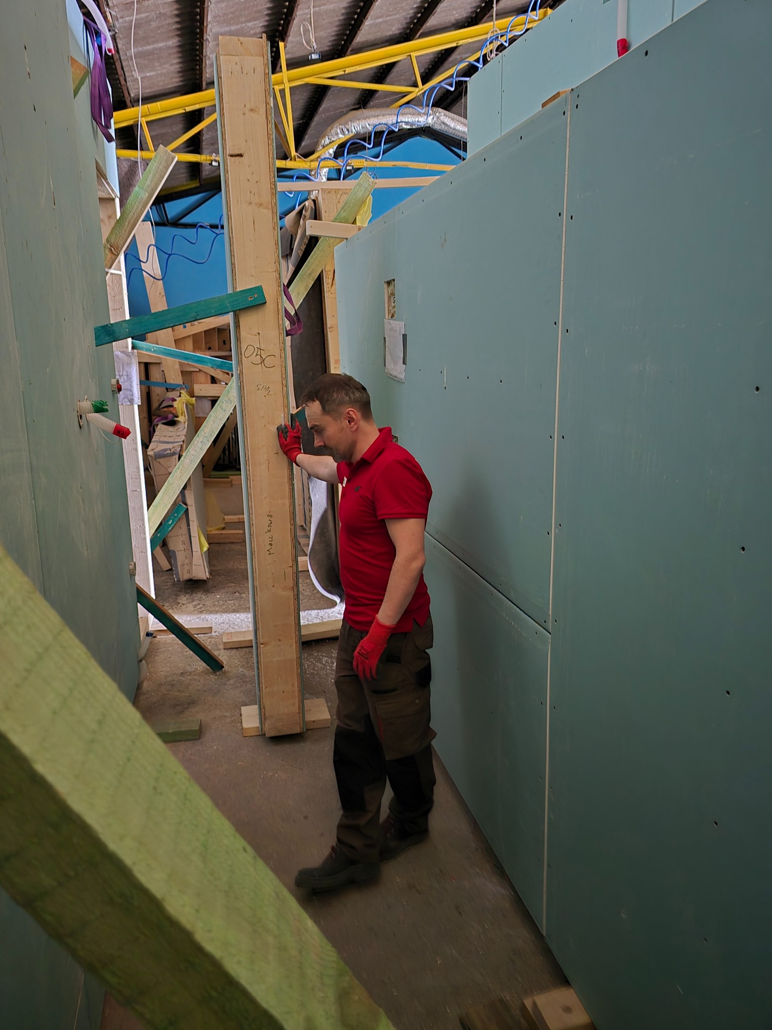 Wnętrze w trakcie budowy: mężczyzna w czerwonej koszulce przy drewnianej konstrukcji, widoczne płyty gipsowe i elementy szkieletu budynku. Oświetlenie sufitowe.