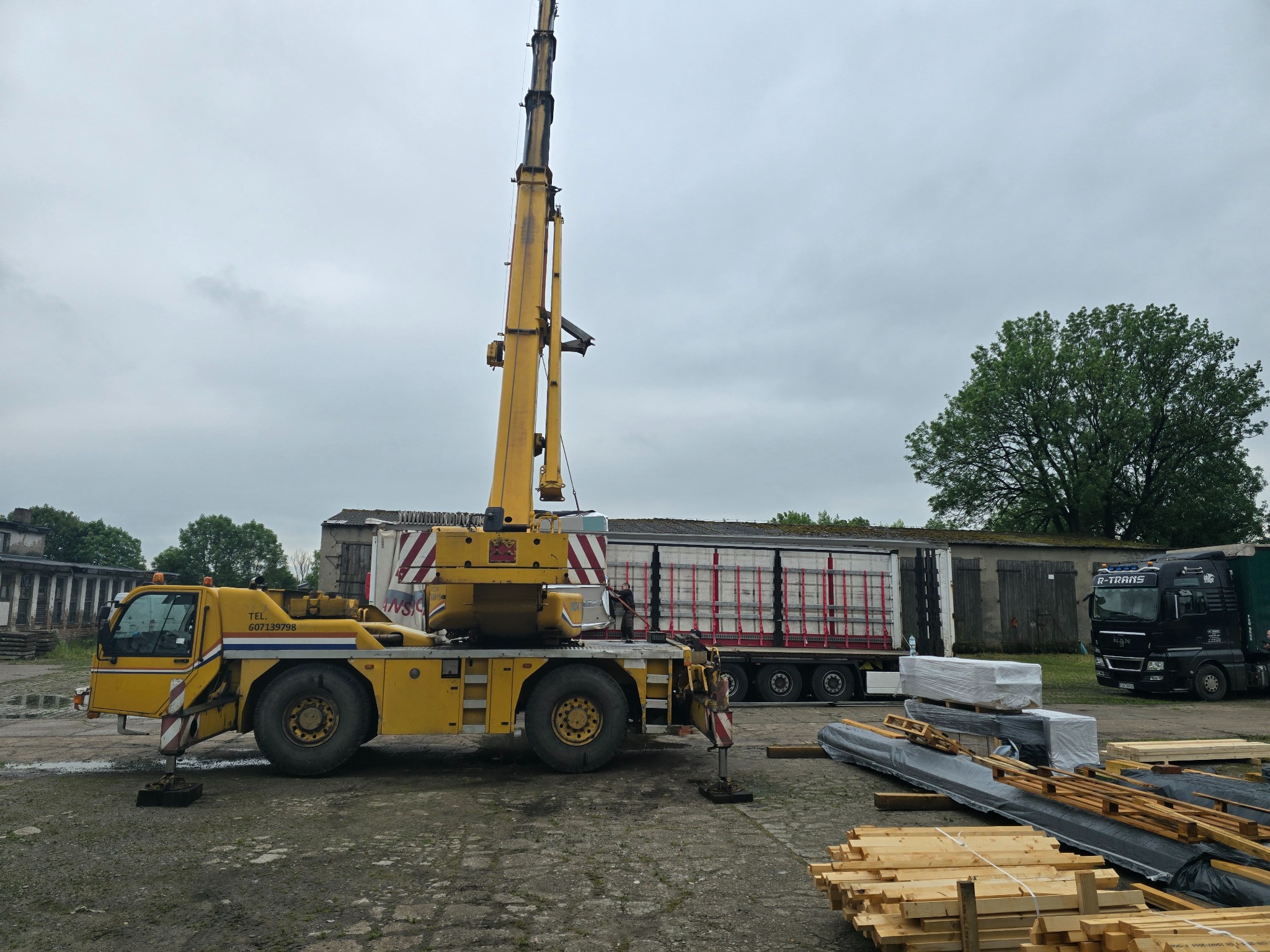 Żółty dźwig teleskopowy na placu budowy, obok leżą drewniane elementy konstrukcyjne i ciężarówka. W tle budynek i drzewo pod pochmurnym niebem.