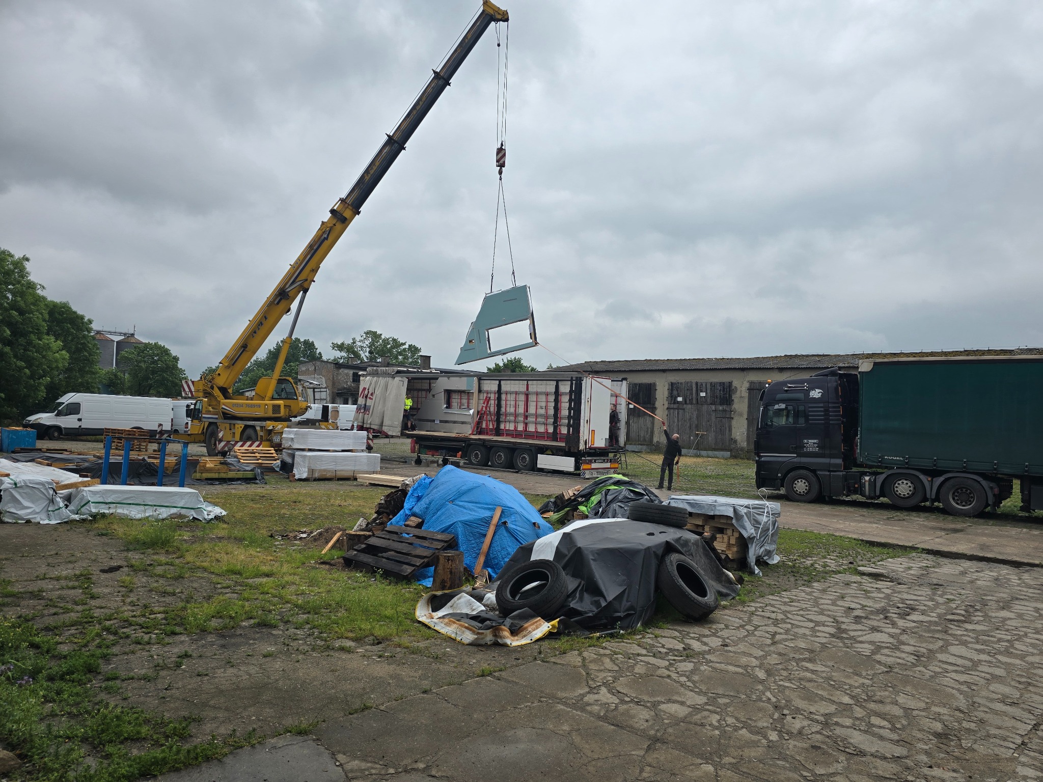 Żuraw przenosi element domu modułowego nad ciężarówką. Widoczne materiały budowlane i pracownik. Pochmurne niebo w tle.
