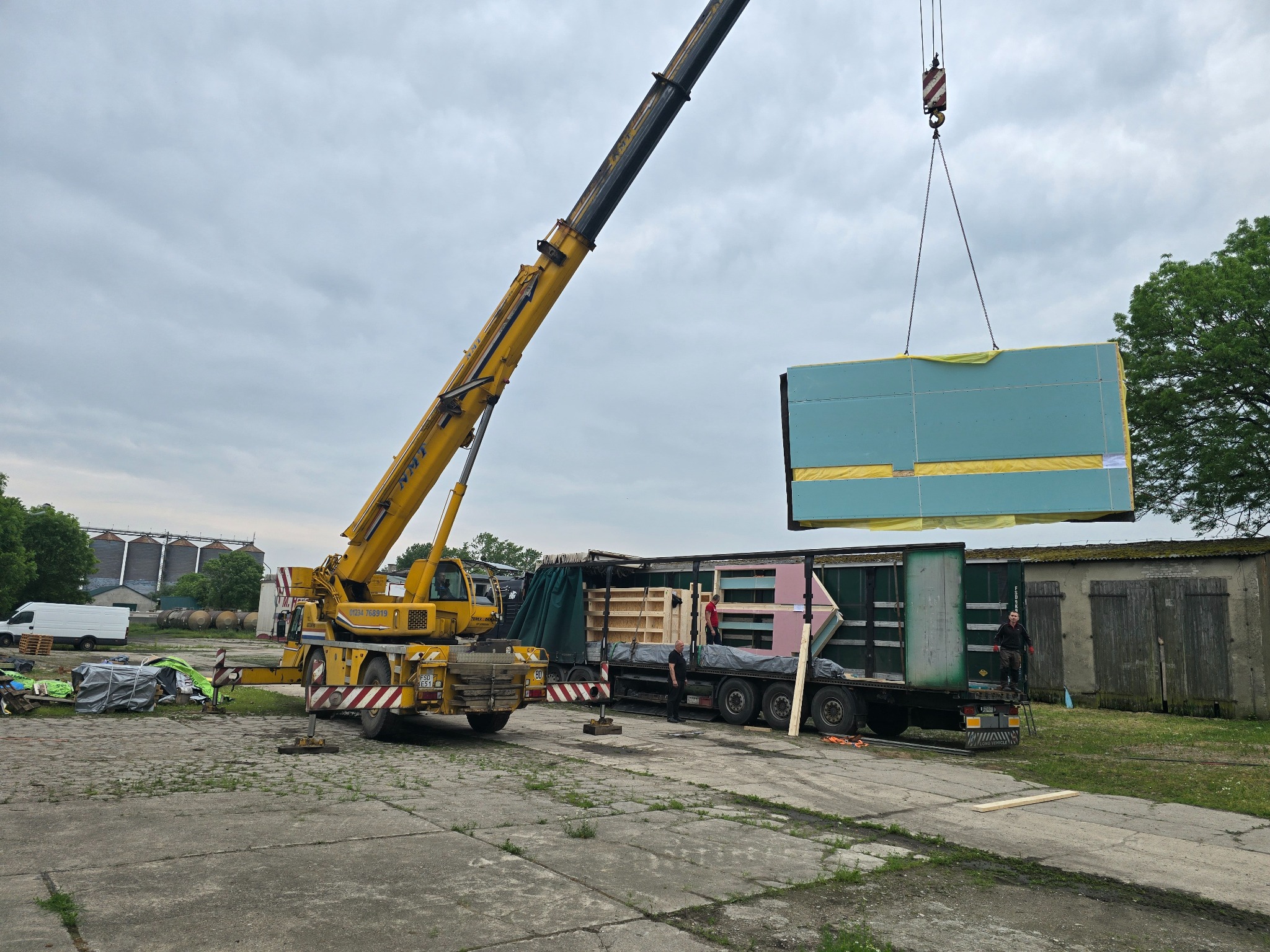 Żuraw przenosi gotowy moduł z niebieską płytą gipsową na ciężarówkę. Widoczni pracownicy i elementy konstrukcyjne. Montaż modułu na placu budowy.