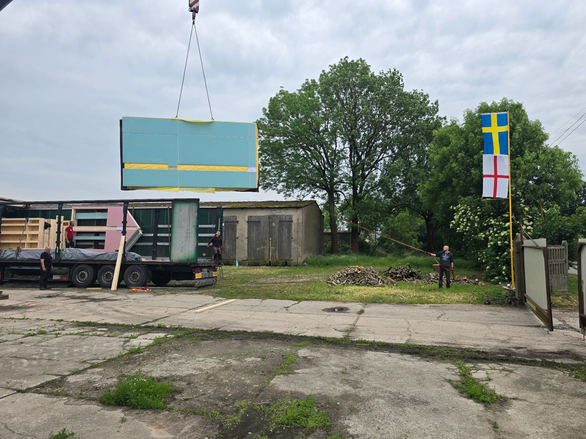 Montaż modułu domu z użyciem dźwigu. Widoczny transport modułów na ciężarówce i flaga Szwecji. Etap budowy z elementami prefabrykowanymi.