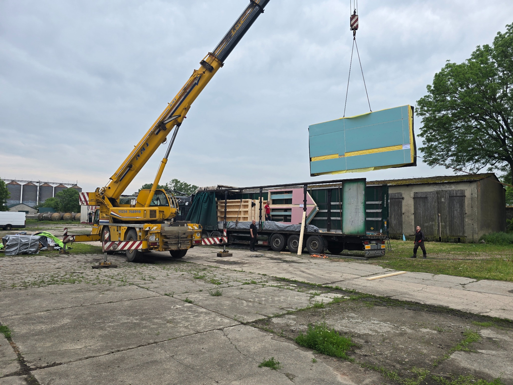 Żuraw przenosi gotowy moduł z niebieską elewacją na ciężarówkę. Widoczni pracownicy i elementy konstrukcyjne. Etap budowy domu.