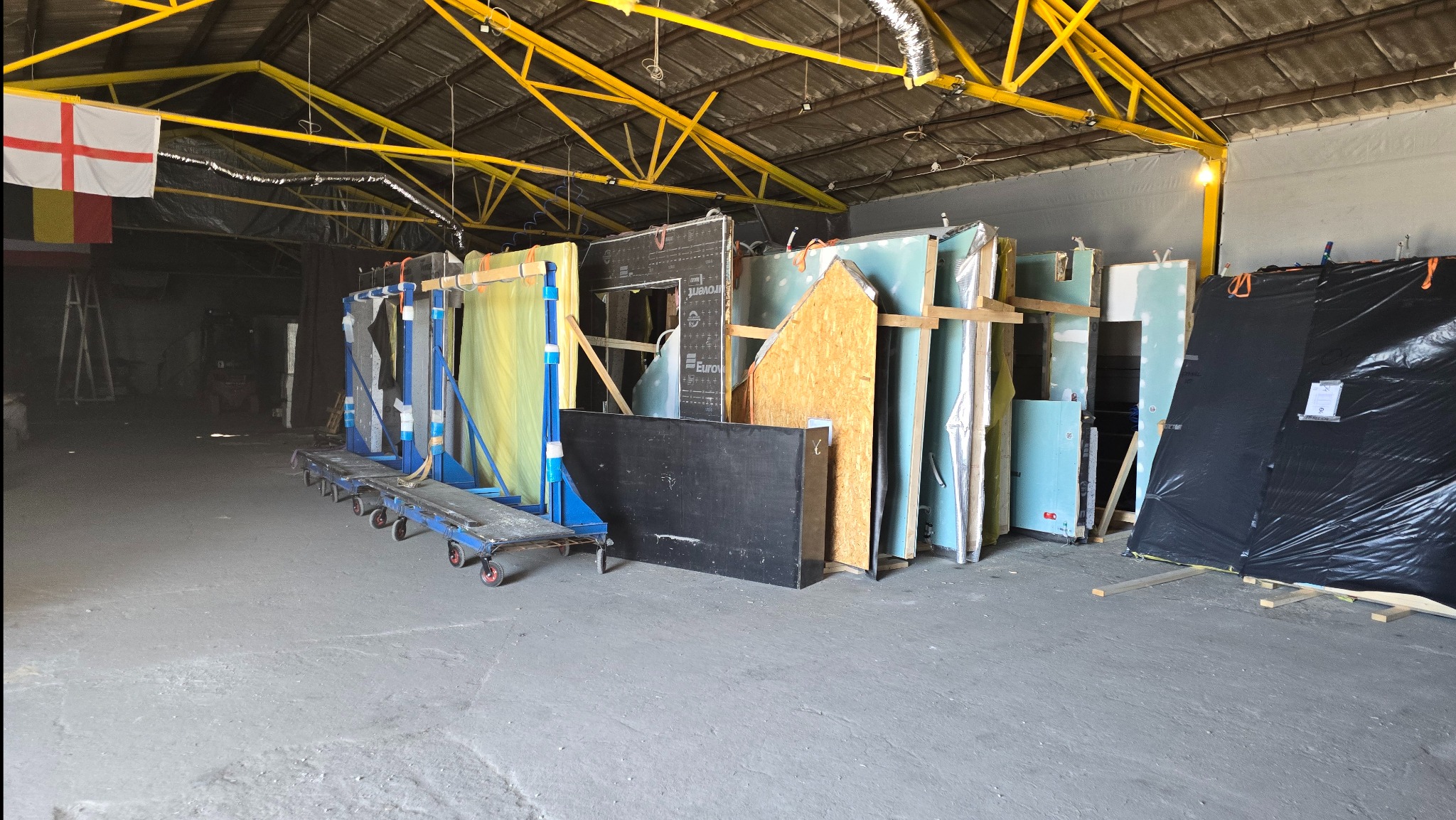 Elementy konstrukcyjne, płyty gipsowe i drewniane, przygotowane do budowy domu, składowane w hali z żółtą konstrukcją dachu i flagami w tle.