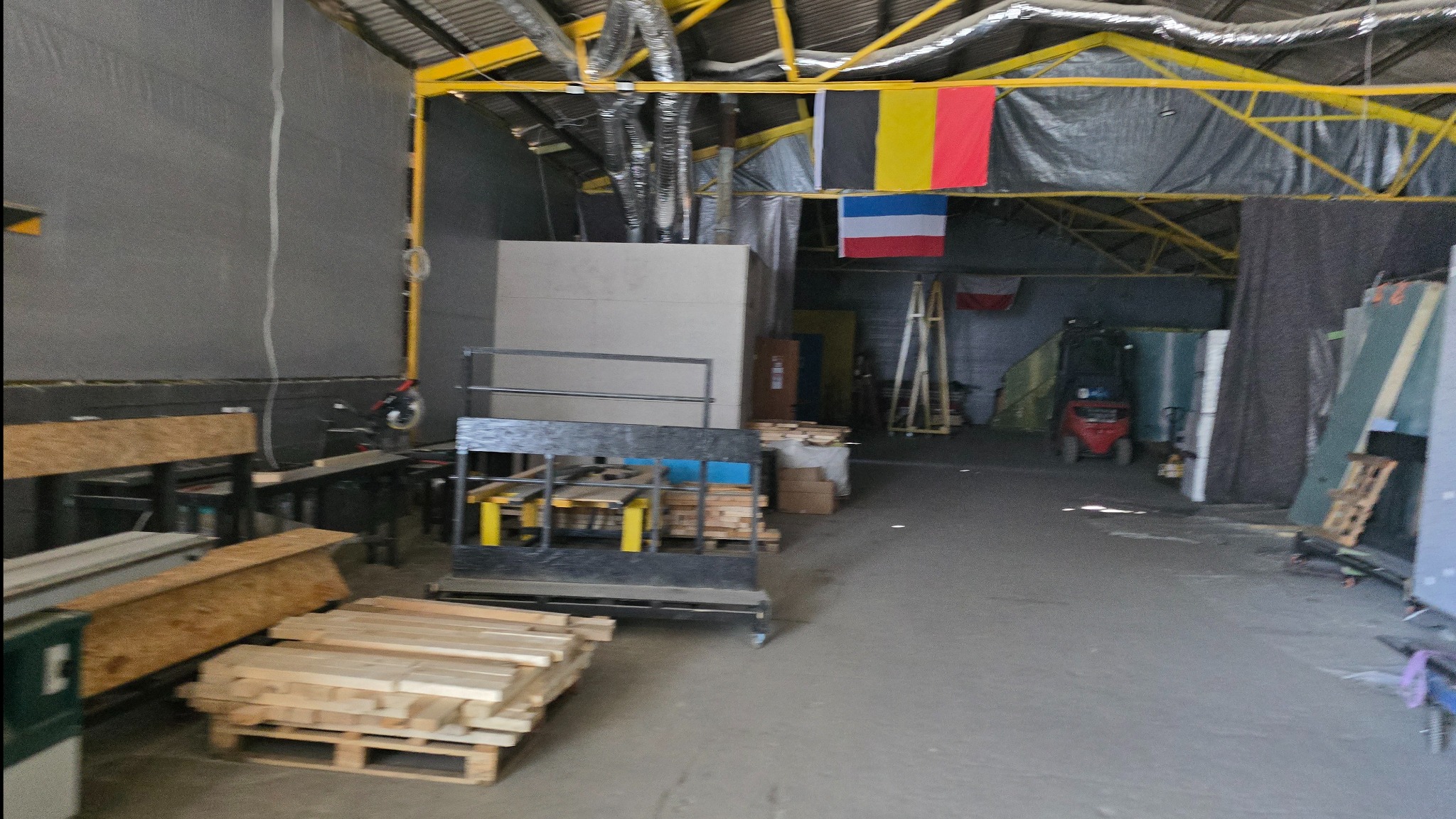 Wnętrze hali produkcyjnej z materiałami budowlanymi, w tle widoczne flagi i wózek widłowy. Elementy konstrukcyjne w kolorze żółtym.