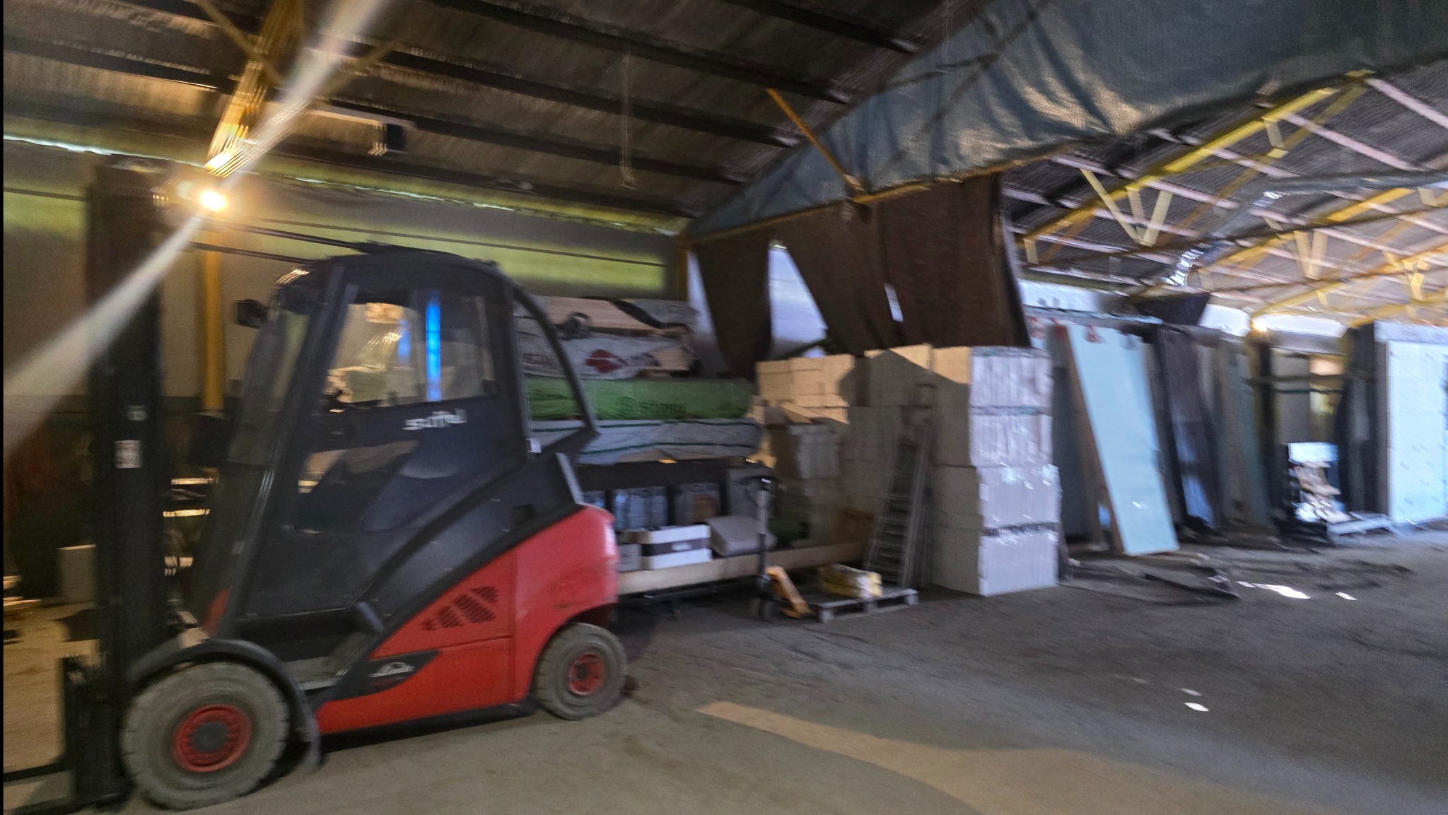 Wnętrze magazynu z wózkiem widłowym Linde, paletami materiałów budowlanych i płytami gipsowymi pod zadaszeniem z metalową konstrukcją.