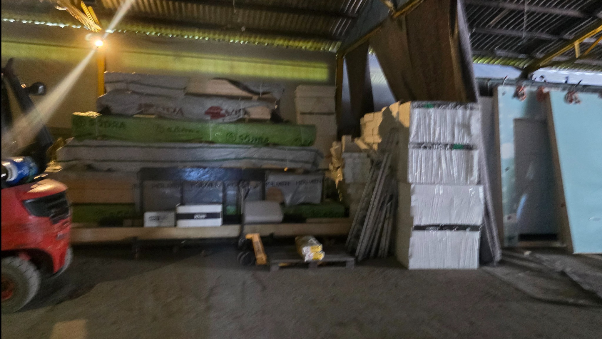 Magazyn materiałów budowlanych: płyty izolacyjne, drewno konstrukcyjne i inne elementy składowane w hali z blachy falistej, oświetlone sztucznym światłem.