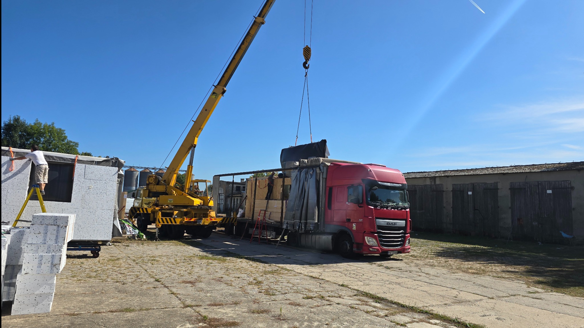 Transport modułu domu z użyciem dźwigu i ciężarówki DAF. Pracownik na drabinie przy module. Słoneczny dzień, błękitne niebo. Widoczne elementy konstrukcyjne.