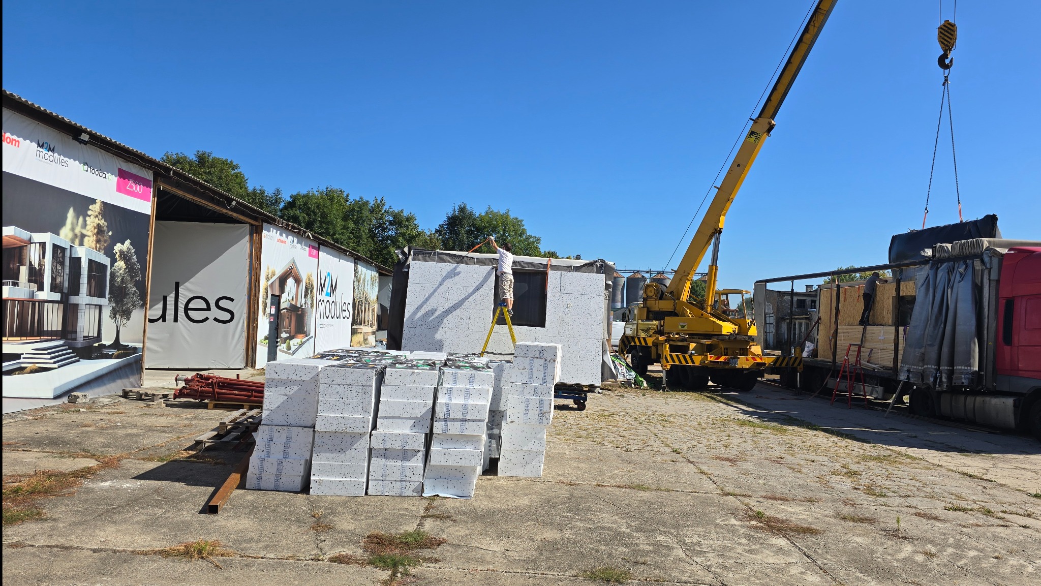 Montaż modułu domu z białych paneli na placu budowy, widoczny dźwig i materiały izolacyjne. Pracownik na drabinie, w tle budynki z reklamami.