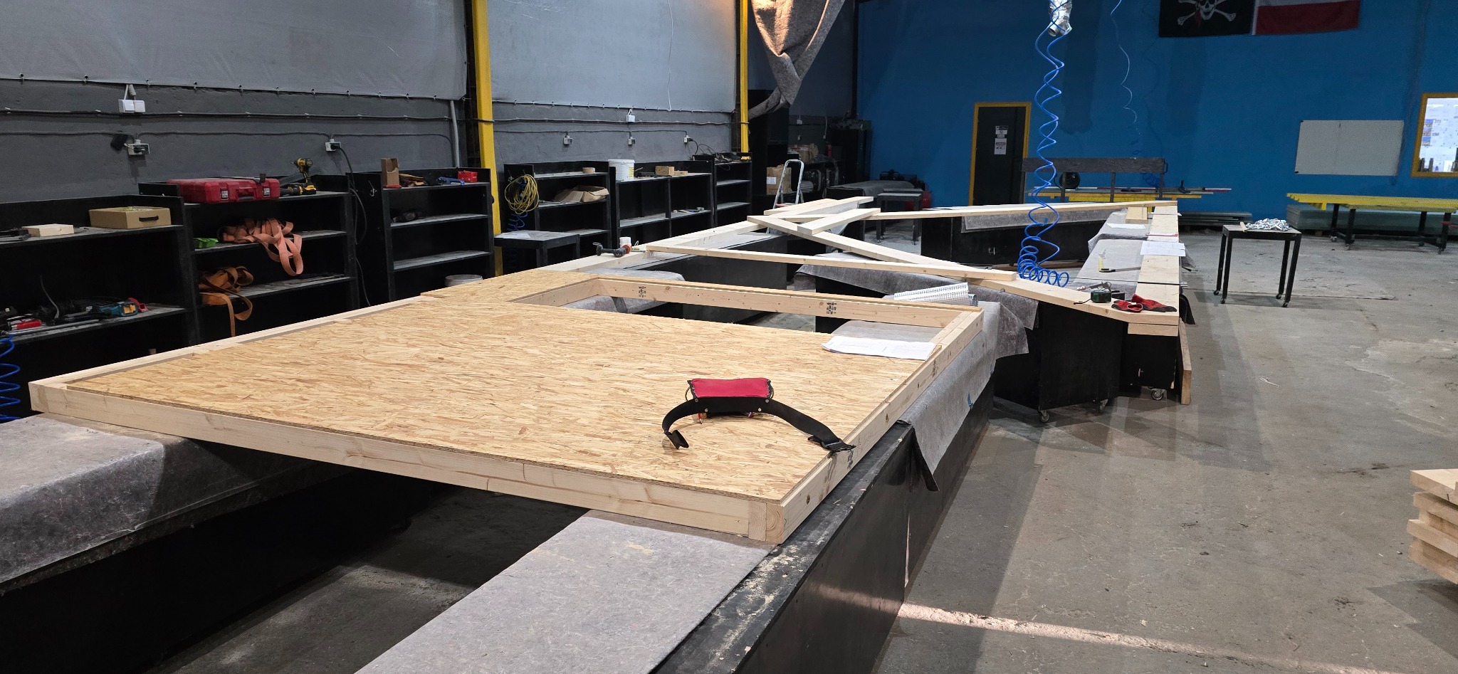 Wnętrze hali produkcyjnej z elementami konstrukcyjnymi ścian z płyty OSB i drewna, przygotowywanymi do budowy domu. Widoczne narzędzia i materiały budowlane.