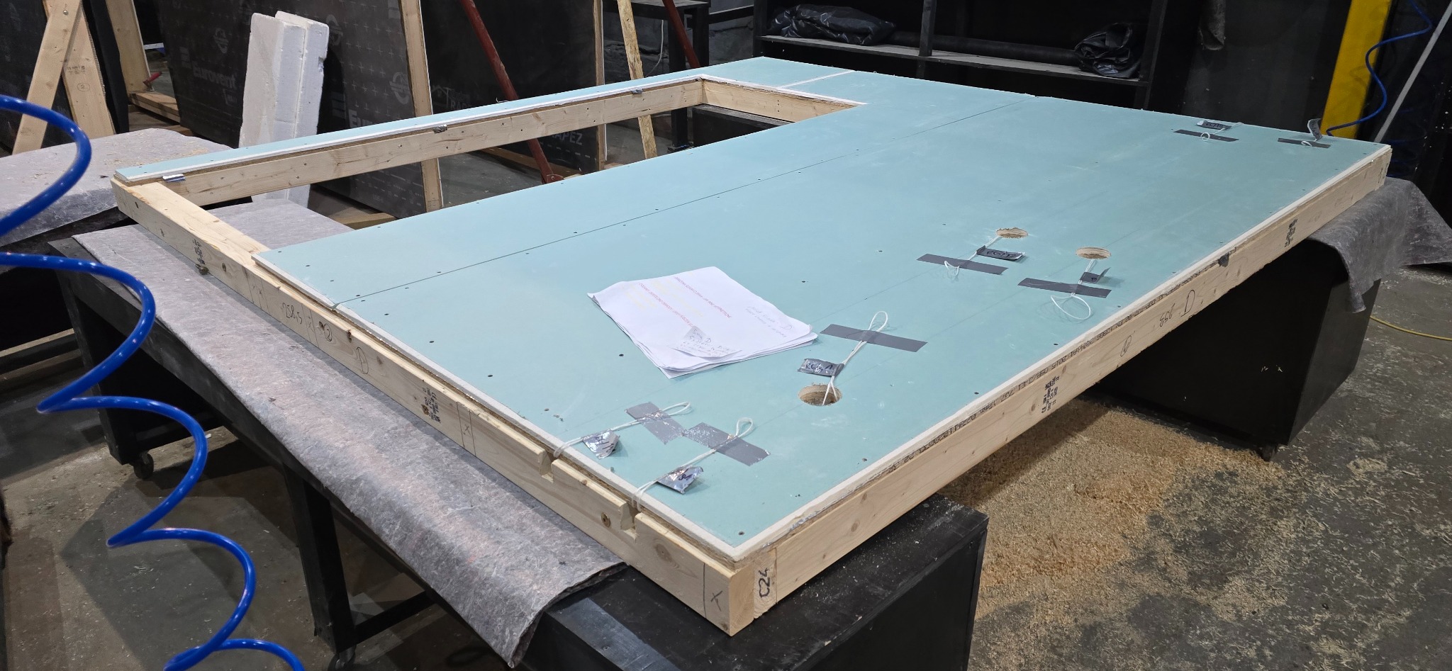 Wnętrze modułu domu w trakcie budowy: drewniana rama z płytą gipsowo-kartonową, widoczne otwory i przewody, dokumentacja techniczna na powierzchni.