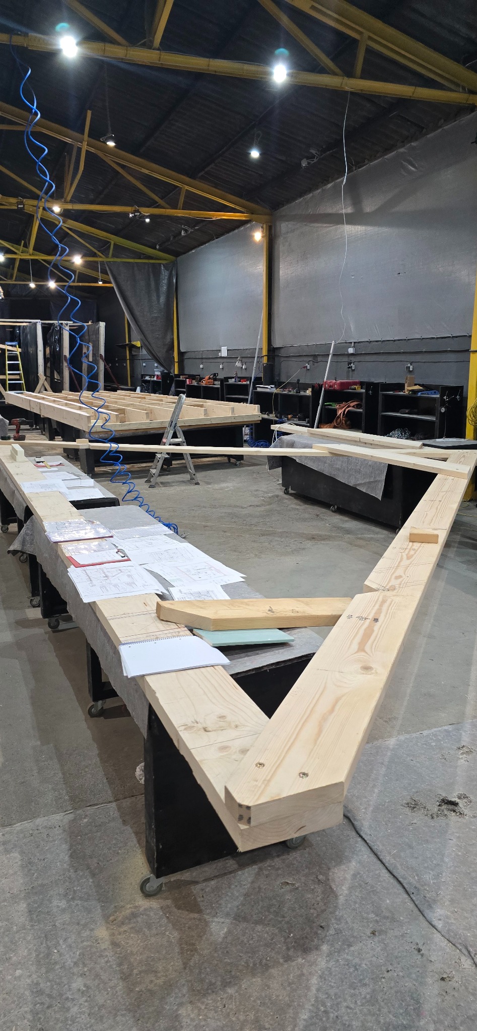 Wnętrze hali produkcyjnej z elementami konstrukcyjnymi z jasnego drewna, przygotowanymi do budowy. Widoczne plany i notatki na stole roboczym.