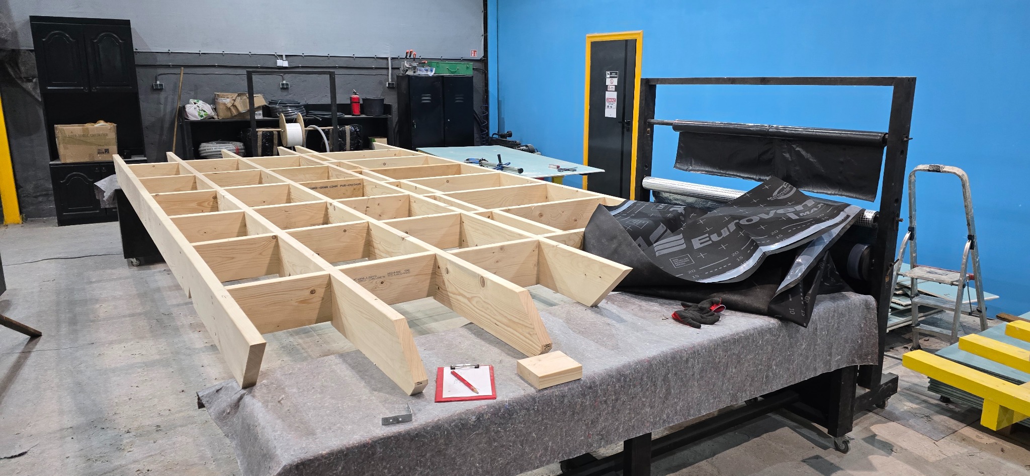 Konstrukcja szkieletowa z drewna na stole warsztatowym, widoczne materiały izolacyjne i narzędzia. Niebieska ściana w tle. Etap budowy domu.