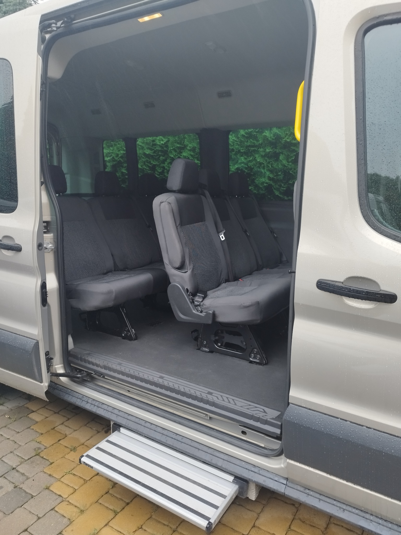 Wnętrze busa z wysuwanym stopniem ułatwiającym wsiadanie. Widoczne fotele z pasami bezpieczeństwa i zielone tło za oknami. Zewnętrzna karoseria w kolorze beżowym.