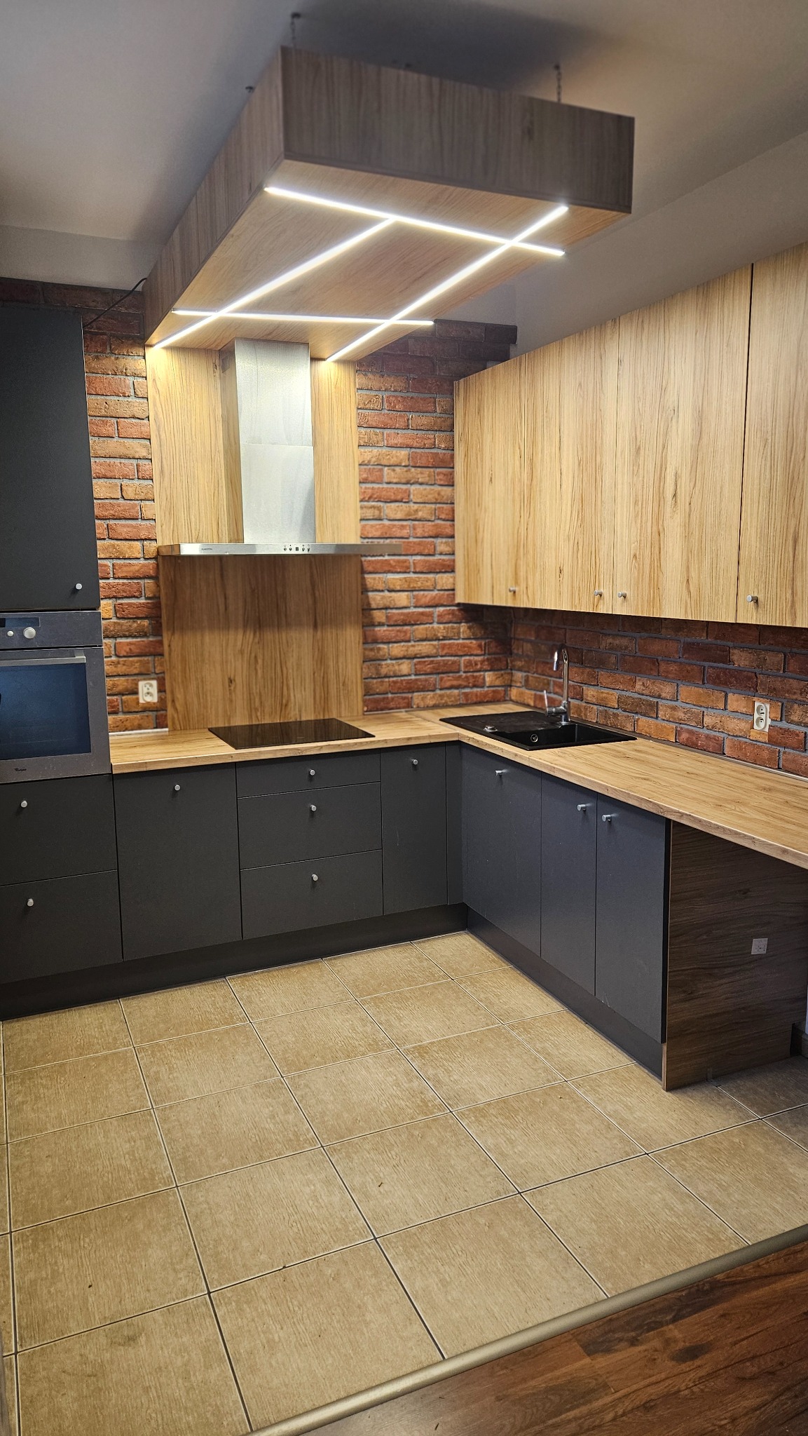 Kątowe ujęcie szaro-drewnianej kuchni z ceglaną ścianą. Górne szafki w kolorze drewna, dolne szare. Nad wyspą kuchenną sufit z podświetleniem LED.