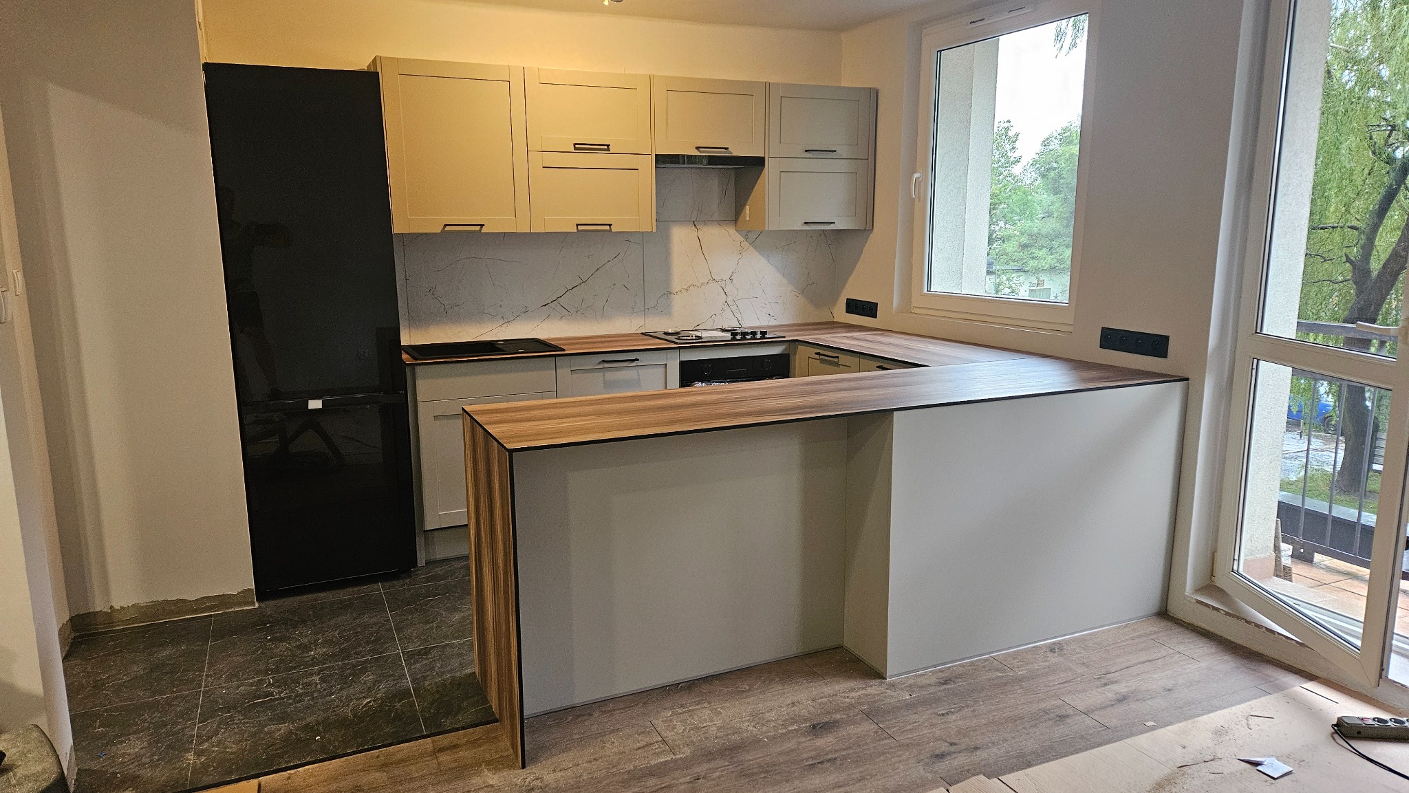 Jasna kuchnia z wyspą i czarną lodówką, widok z boku. Marmurowy backsplash i drewniany blat dodają elegancji. Duże okno wpuszcza naturalne światło.