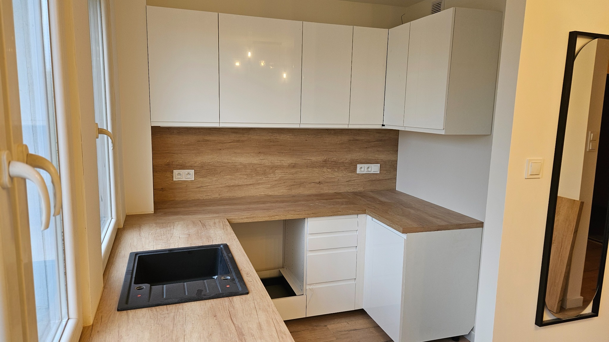 Nowoczesna kuchnia z białymi, lakierowanymi szafkami i drewnianym blatem. Uwagę przyciąga czarny zlew wkomponowany w jasny blat. Całość doświetlona naturalnym światłem.