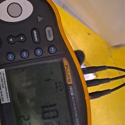 VSpark Volodymyr Semenikhin - Miernik Fluke 1653B Multifunction Tester z podłączonymi przewodami pomiarowymi, wskazujący wartości pomiarowe na wyświetlaczu. Widoczny fragment żółtej powierzchni.
