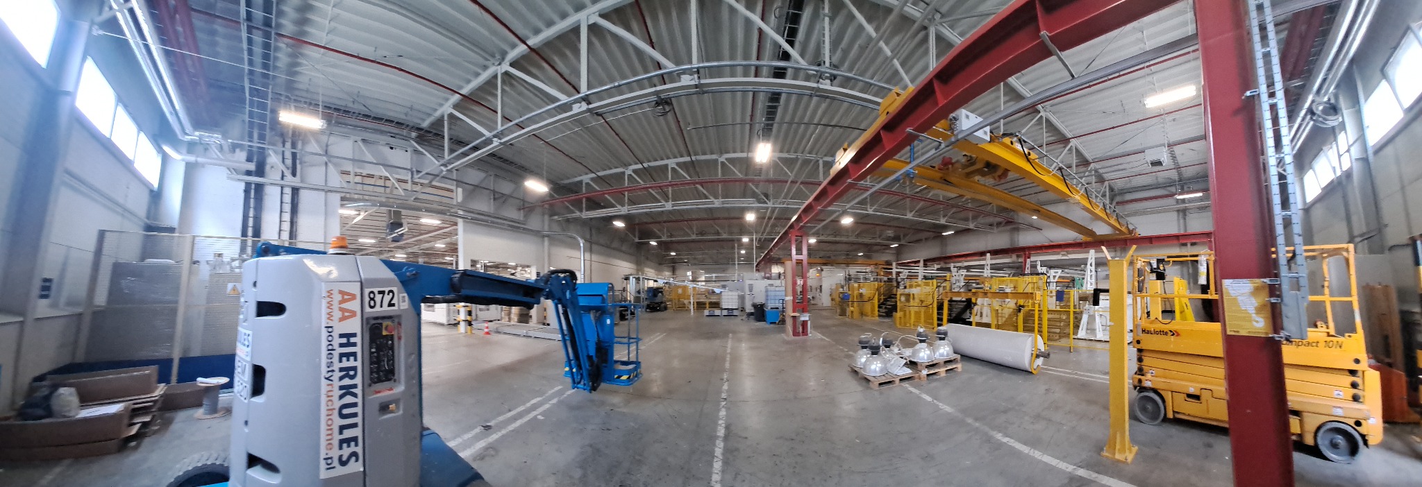 Szeroki kadr hali produkcyjnej z podnośnikami, widoczne metalowe konstrukcje sufitu i oświetlenie przemysłowe. Paleta z lampami na podłodze.