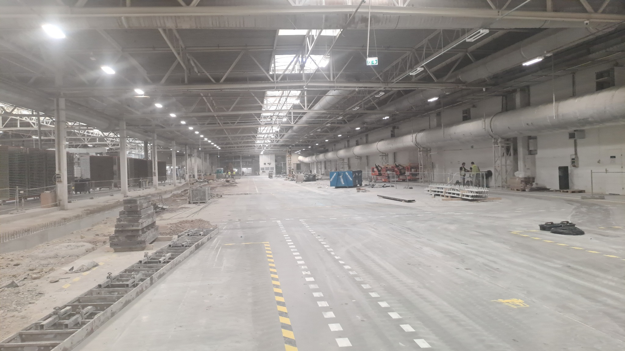 Wnętrze hali przemysłowej w trakcie budowy: widoczne instalacje pod sufitem, ekipa budowlana w oddali oraz elementy konstrukcyjne na pierwszym planie.