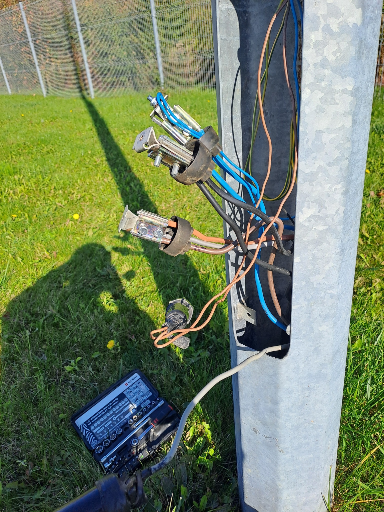 Otwarta skrzynka elektryczna na słupie z wystającymi przewodami i zestawem narzędzi Würth na trawie, sugerująca prace elektryczne w otoczeniu zieleni.
