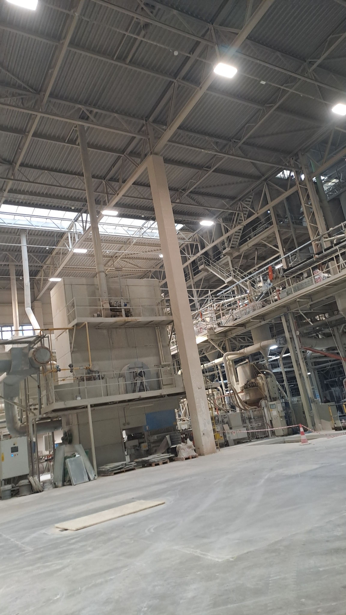 Wnętrze hali przemysłowej z metalową konstrukcją, oświetleniem i instalacjami. Widoczne elementy maszyn i urządzeń. Perspektywa z dołu, ukazująca rozległość przestrzeni.