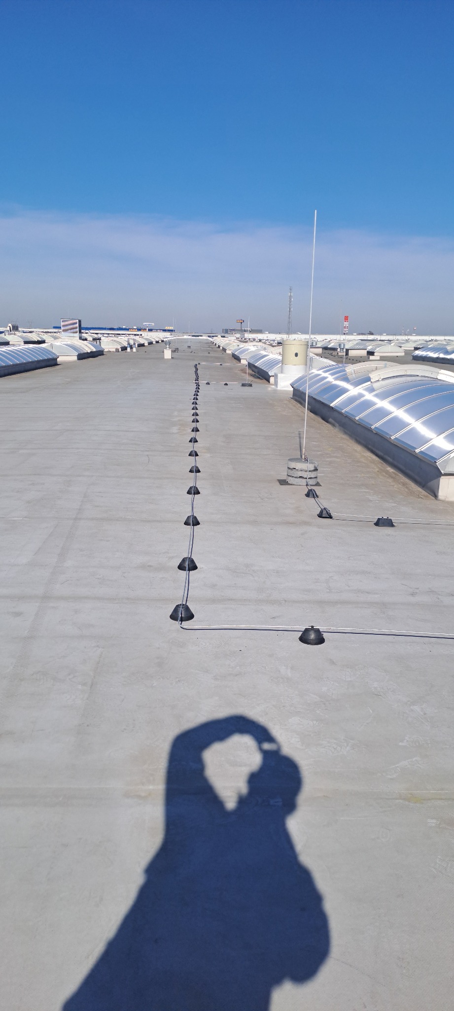 Instalacja odgromowa na dachu budynku. Widoczne elementy mocujące przewody i zwody pionowe. W tle świetliki dachowe i shadow autora zdjęcia.