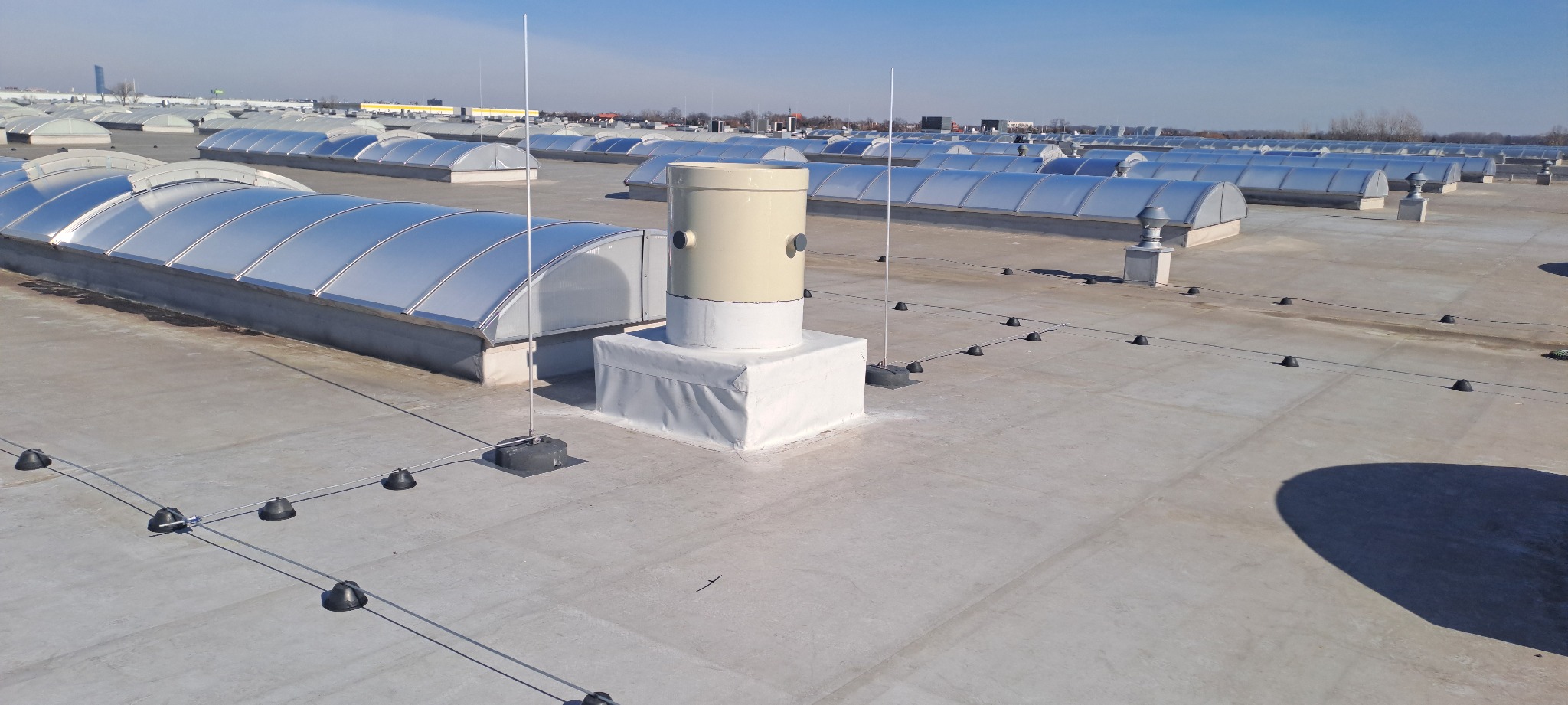 Instalacja odgromowa na dachu budynku z świetlikami dachowymi, widoczne elementy systemu, przewody i uchwyty, na tle niebieskiego nieba.