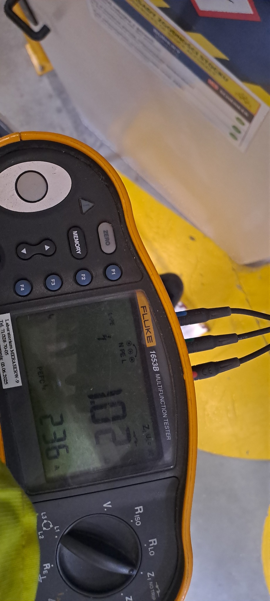 Miernik Fluke 1653B Multifunction Tester z podłączonymi przewodami pomiarowymi, wskazujący wartości pomiarowe na wyświetlaczu. Widoczny fragment żółtej powierzchni.