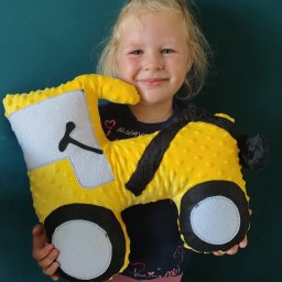 Iwona Pietrzak Nitką pisane - Uśmiechnięta dziewczynka trzyma żółtą, pluszową poduszkę w kształcie traktora z czarnymi detalami na tle turkusowej ściany. Widoczne szwy i faktura materiału.