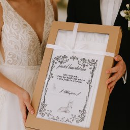 Iwona Pietrzak Nitką pisane - Para młoda prezentuje zapakowany komplet pościeli bawełnianej w ozdobnym pudełku z okienkiem, przewiązany białą wstążką. Widoczny rysunek sarenki i specyfikacja.