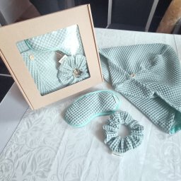 Iwona Pietrzak Nitką pisane - Komplet pościeli w pudełku i obok: szlafrok z kapturem, opaska na oczy i gumka do włosów z waflowej tkaniny w odcieniach błękitu, na białym obrusie.