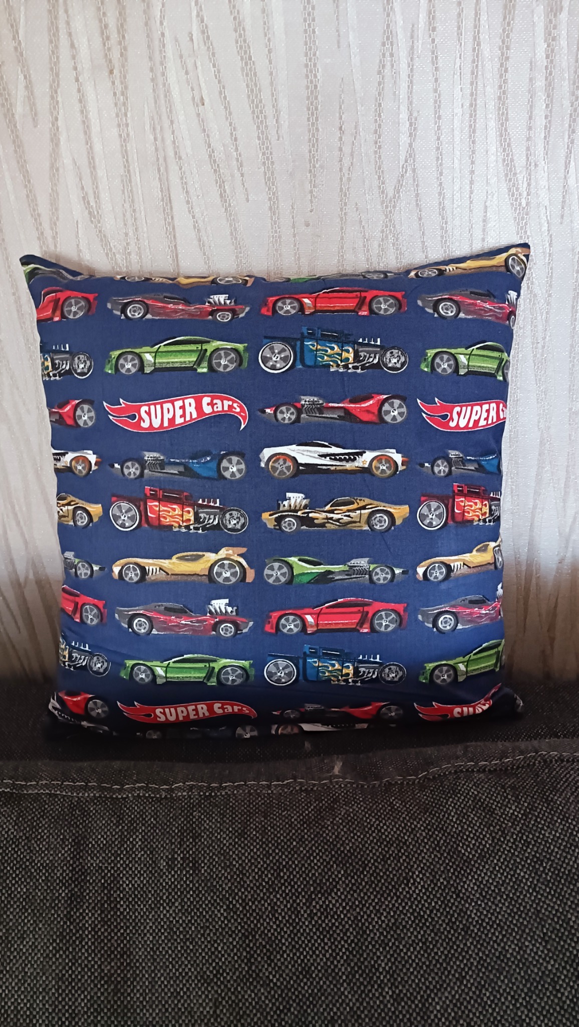 Kwadratowa poduszka dekoracyjna z niebieskiego materiału w kolorowe autka 'Super Cars', leżąca na ciemnoszarym tle. Widoczny fragment białej zasłony w tle.