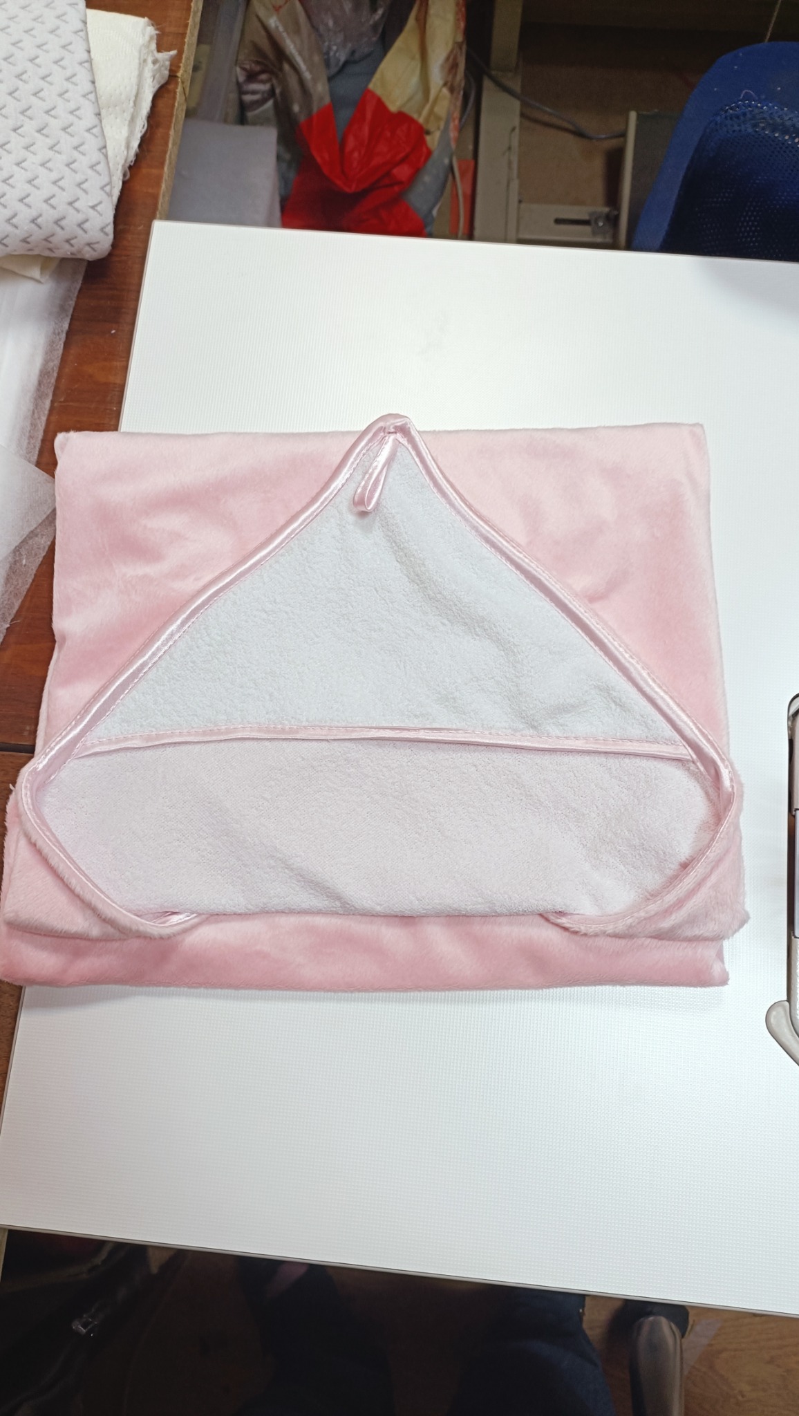 Różowy, miękki ręcznik kąpielowy z kapturem, wykończony białą lamówką, leży złożony na białym blacie. Widoczne inne materiały i elementy wyposażenia szwalni w tle.