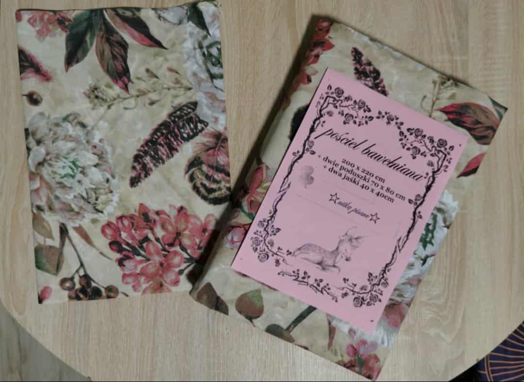 Zestaw pościeli bawełnianej w kwiaty z etykietą informacyjną na drewnianym stole. Widoczne wymiary: 200x220 cm, dwie poduszki 70x80 cm, dwa jaśki 40x40 cm.