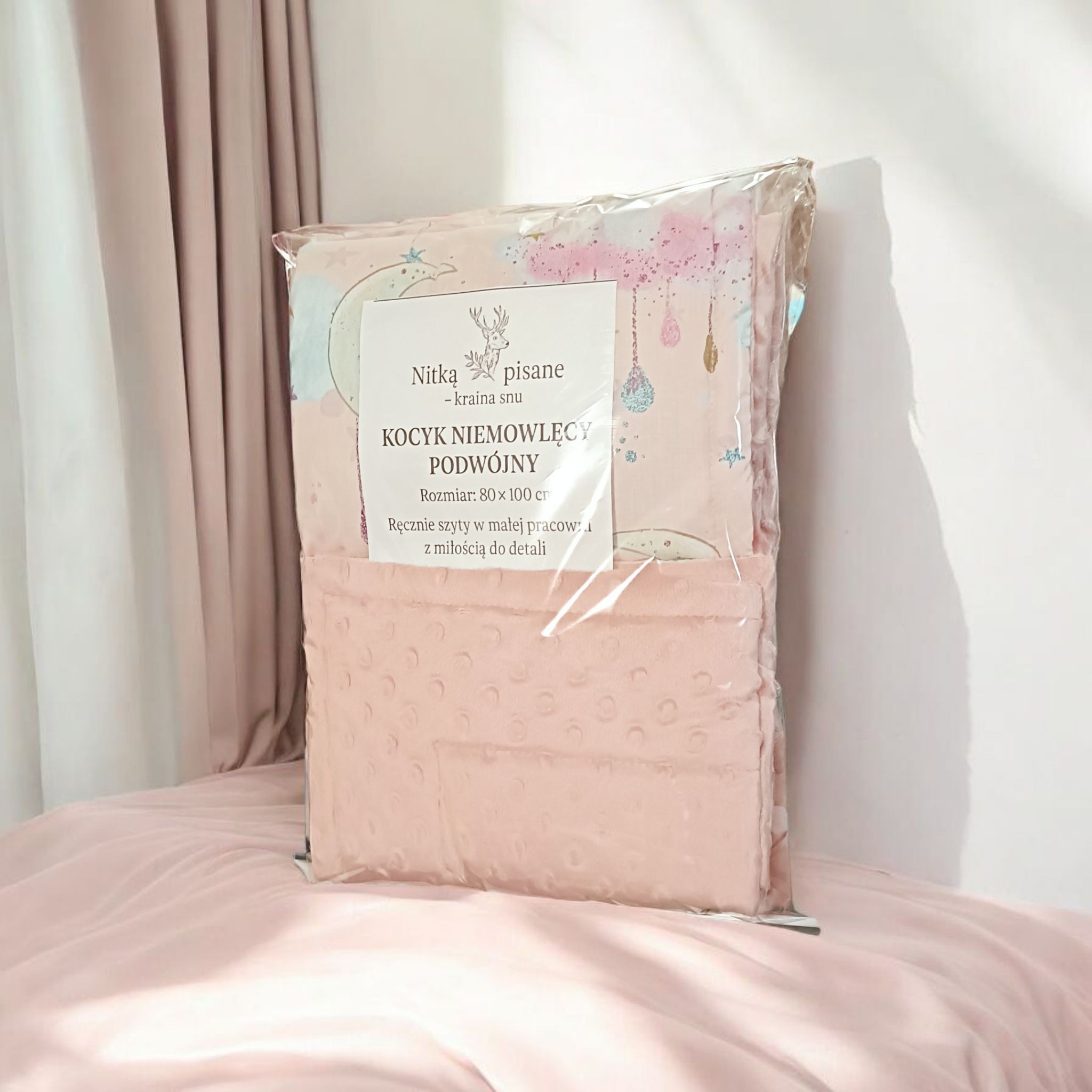 Ręcznie szyty kocyk niemowlęcy 80x100 cm, zapakowany w folię, z metką 'Nitką pisane' z Gniezna, leży na różowej pościeli. Pastelowe kolory, delikatny wzór.