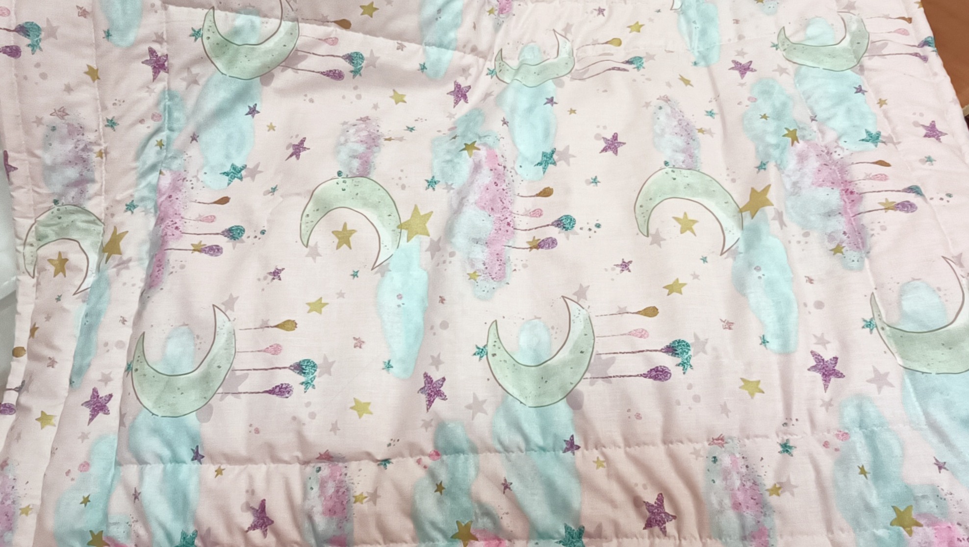 Różowa kołdra dziecięca w księżyce i gwiazdki, pikowana w regularne kwadraty, z bliska. Pastelowe kolory, miękka tkanina, idealna do dziecięcego pokoju.