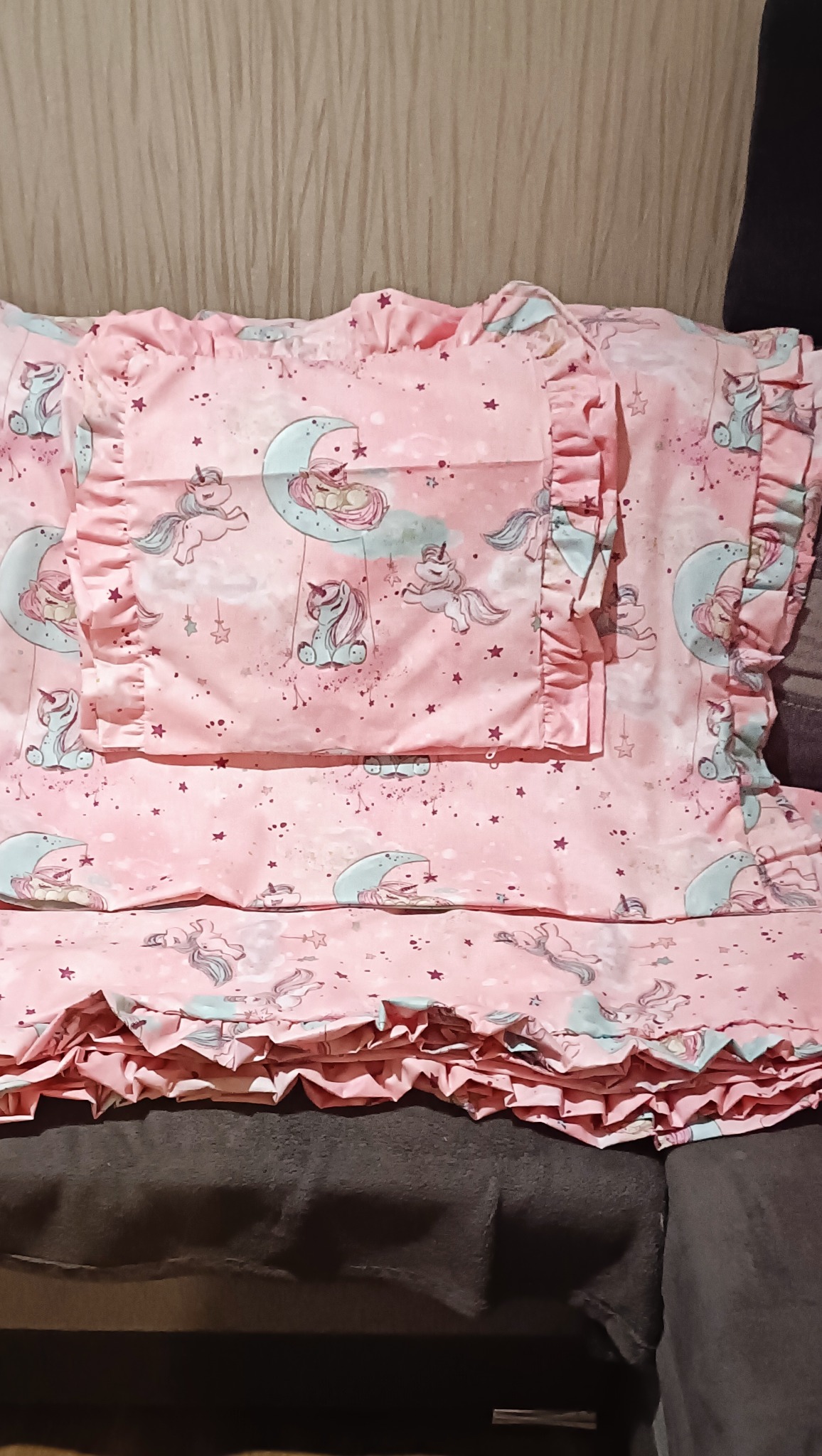 Różowa pościel dziecięca z falbankami i motywem jednorożców na księżycu, leżąca na ciemnej kanapie. Widoczne poduszki i kołdra.