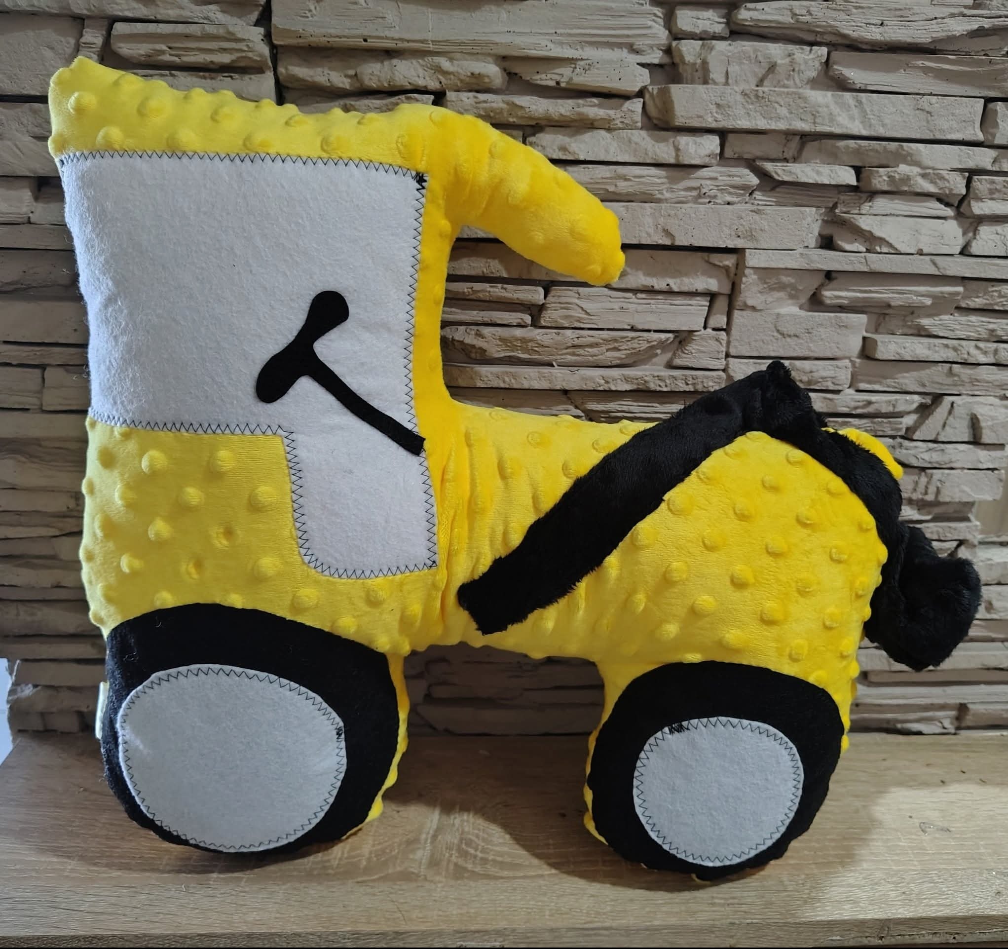 Żółto-biało-czarna, pluszowa poduszka w kształcie traktora, z wyraźnymi szwami, na tle ściany z kamienia dekoracyjnego. Zabawka ręcznie szyta, widoczne detale.