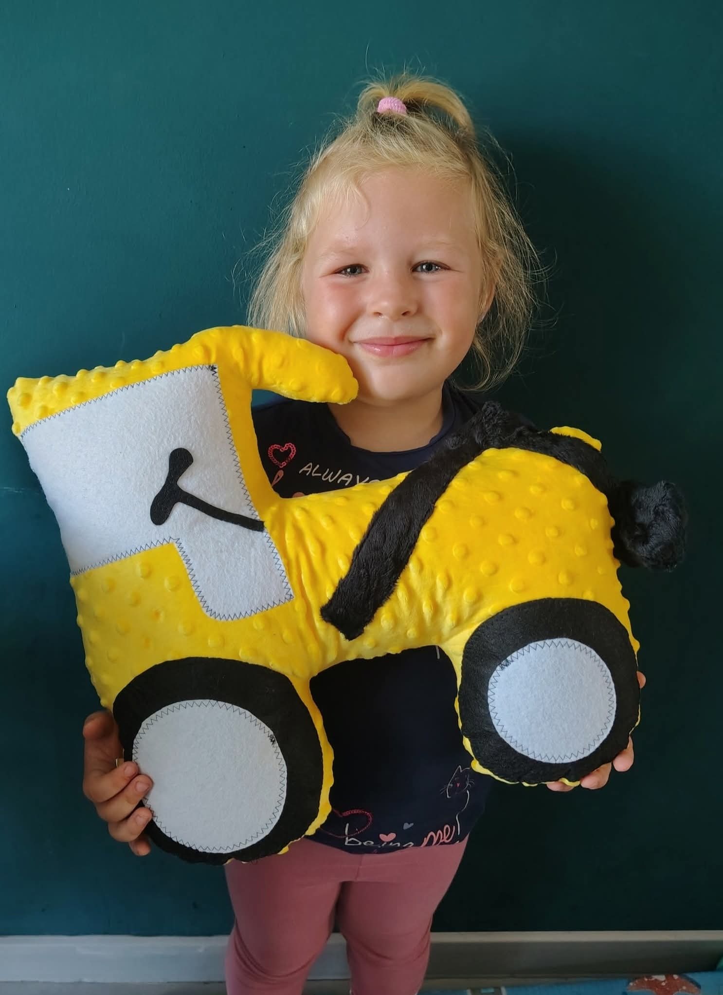 Uśmiechnięta dziewczynka trzyma żółtą, pluszową poduszkę w kształcie traktora z czarnymi detalami na tle turkusowej ściany. Widoczne szwy i faktura materiału.