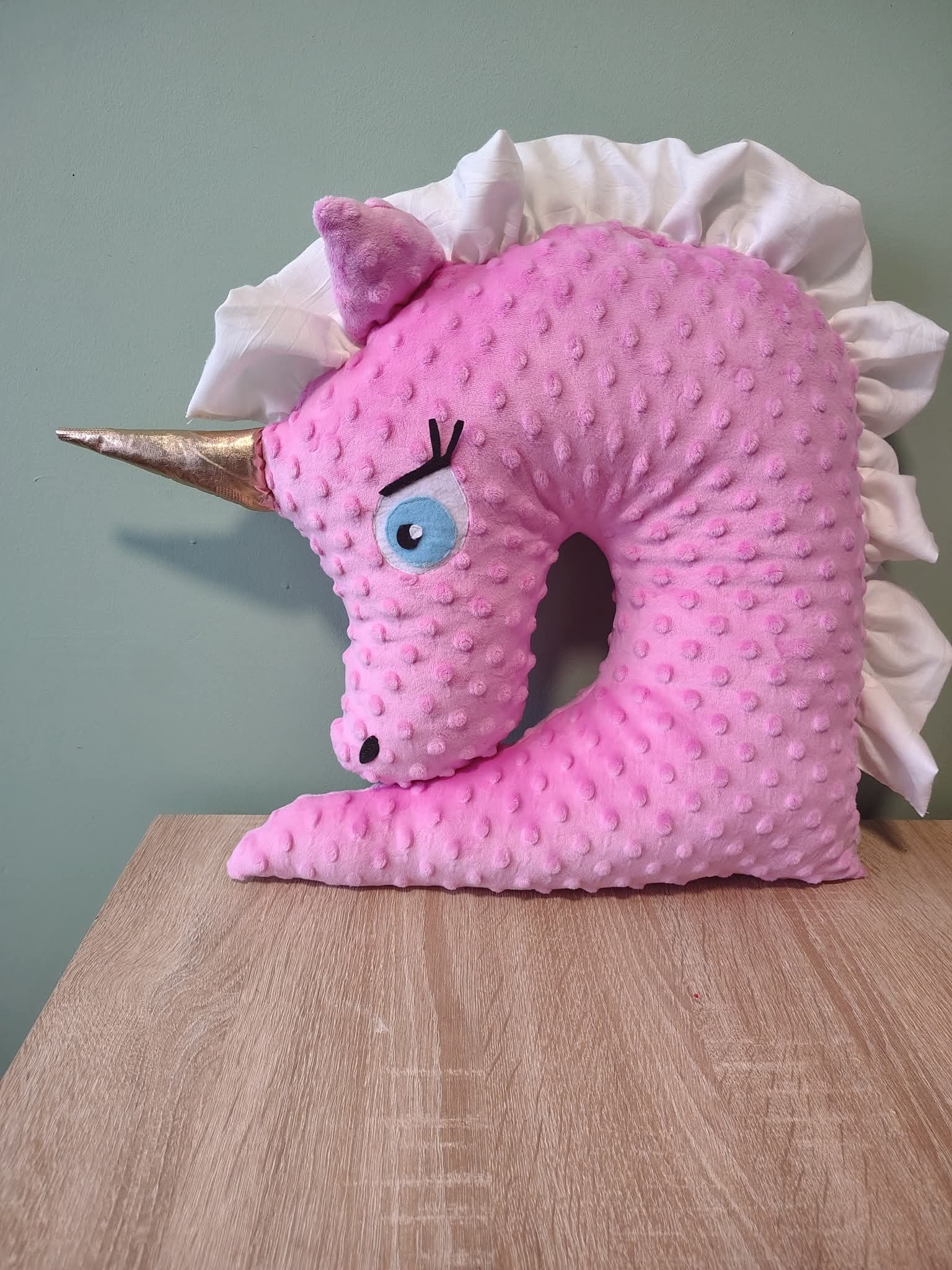 Ręcznie szyta, różowa poduszka w kształcie głowy jednorożca z falbankami, leżąca na drewnianym blacie. Wyraźna faktura materiału i złoty róg.