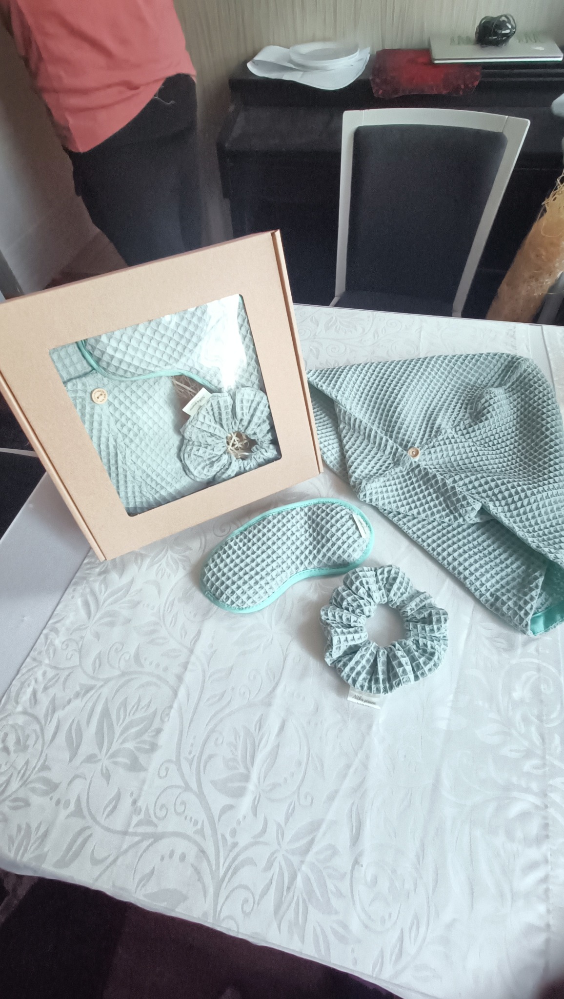 Komplet pościeli w pudełku i obok: szlafrok z kapturem, opaska na oczy i gumka do włosów z waflowej tkaniny w odcieniach błękitu, na białym obrusie.